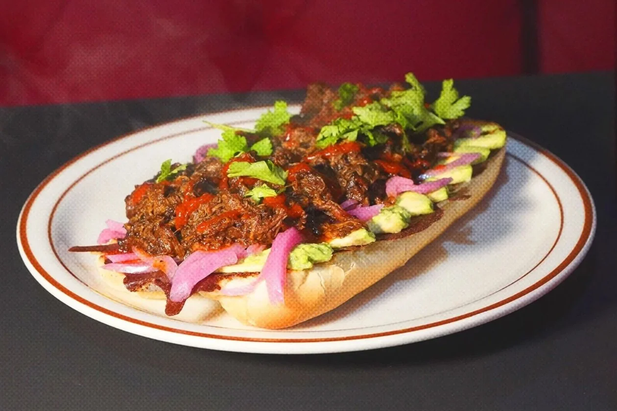 An Italian-style sandwich or hot dog bun filled with seasoned shredded beef, topped with pickled red onions, chopped cilantro, and sliced avocado, served on a white plate with a brown striped rim.