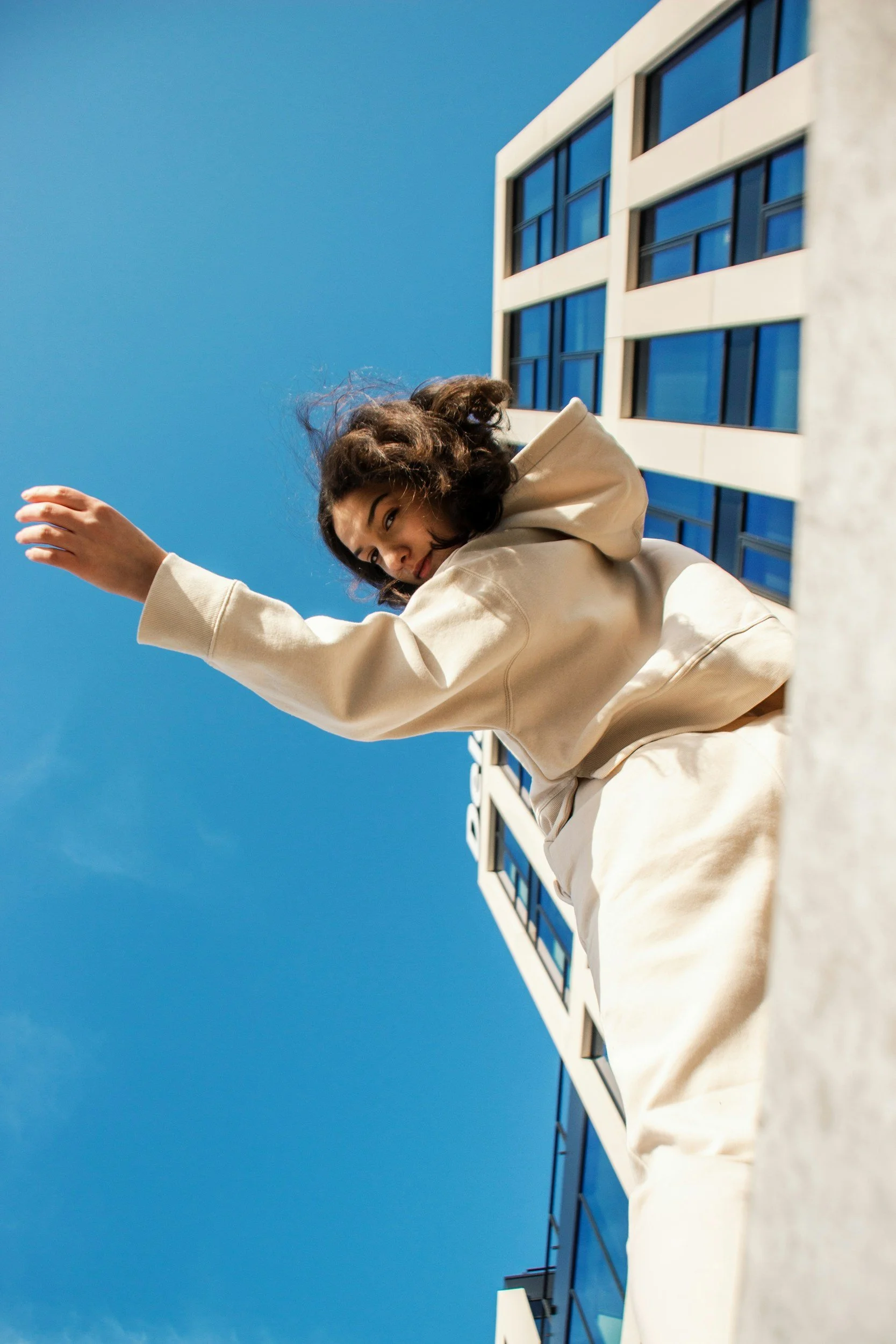A woman with curly hair wearing a beige hoodie and matching pants is reaching out. She is outdoors in front of a modern building with a blue glass facade and white framing, under a clear blue sky.