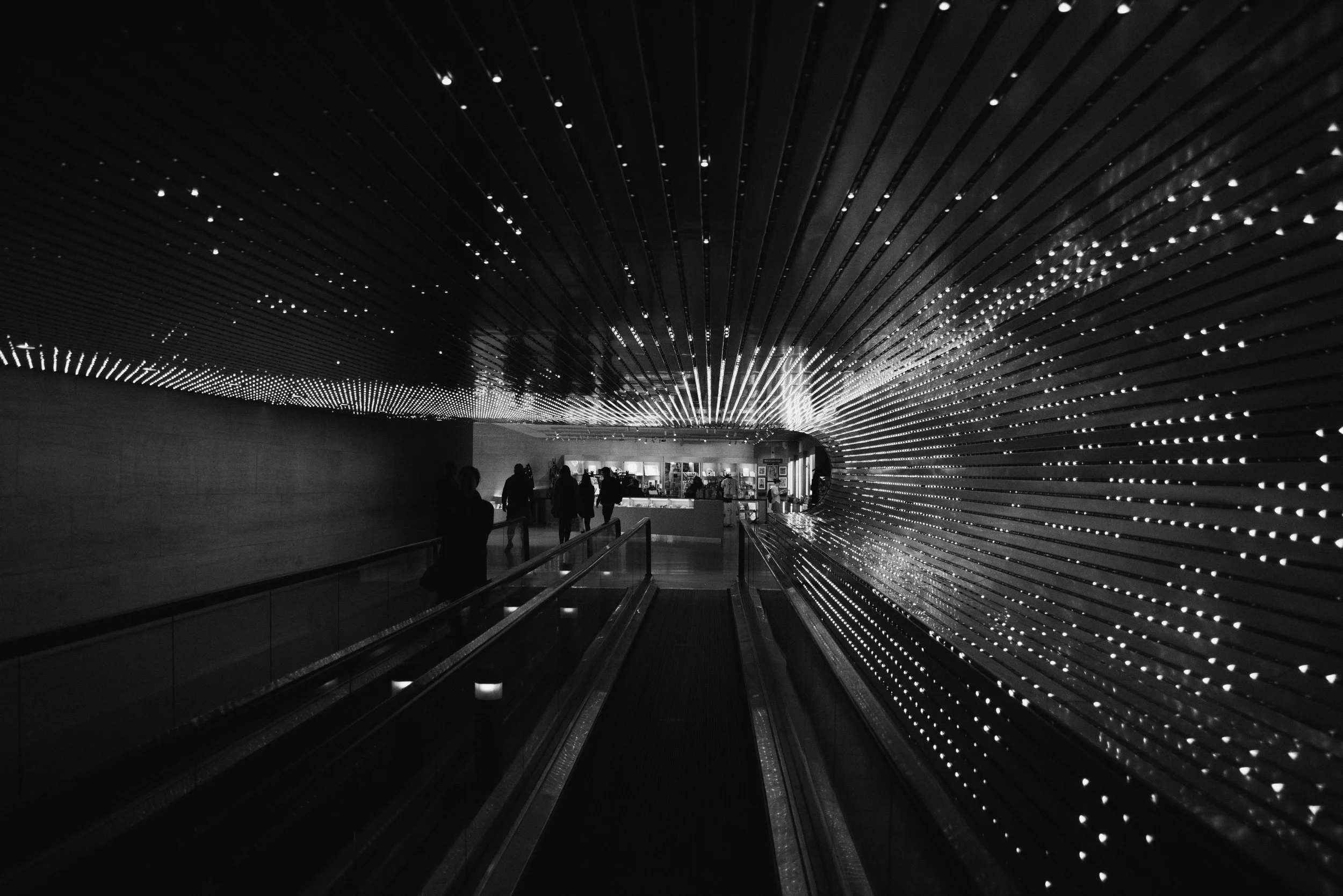 Black and white photo of an underground walkway with a curved wall decorated with numerous small lights, with people walking through.