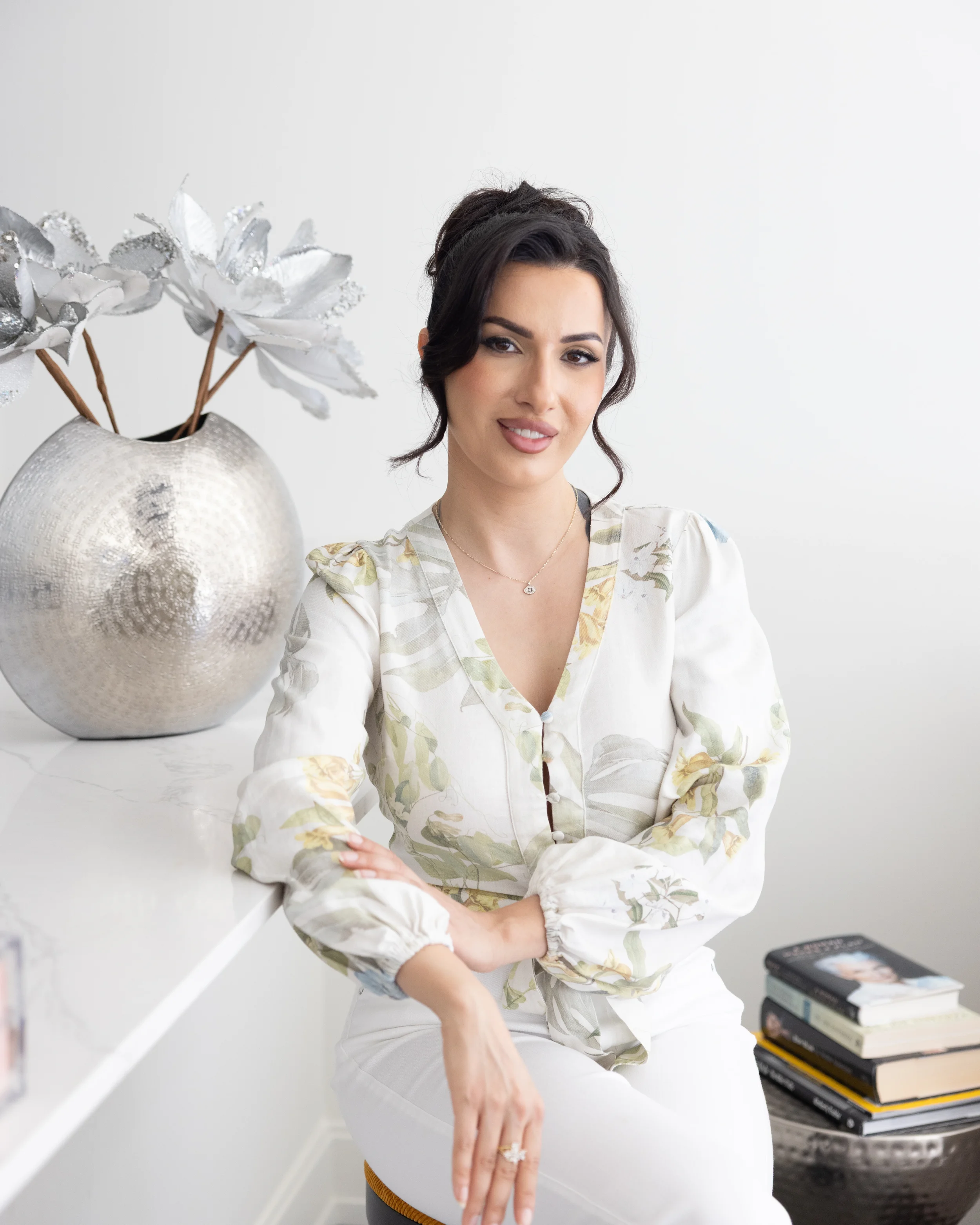A woman with dark hair in a bun, wearing a floral-patterned blouse and white pants, sitting in front of a white wall and a large silver flower vase with decorative white and silver flowers. There is a stack of magazines with a remote on top on a black table in the foreground.