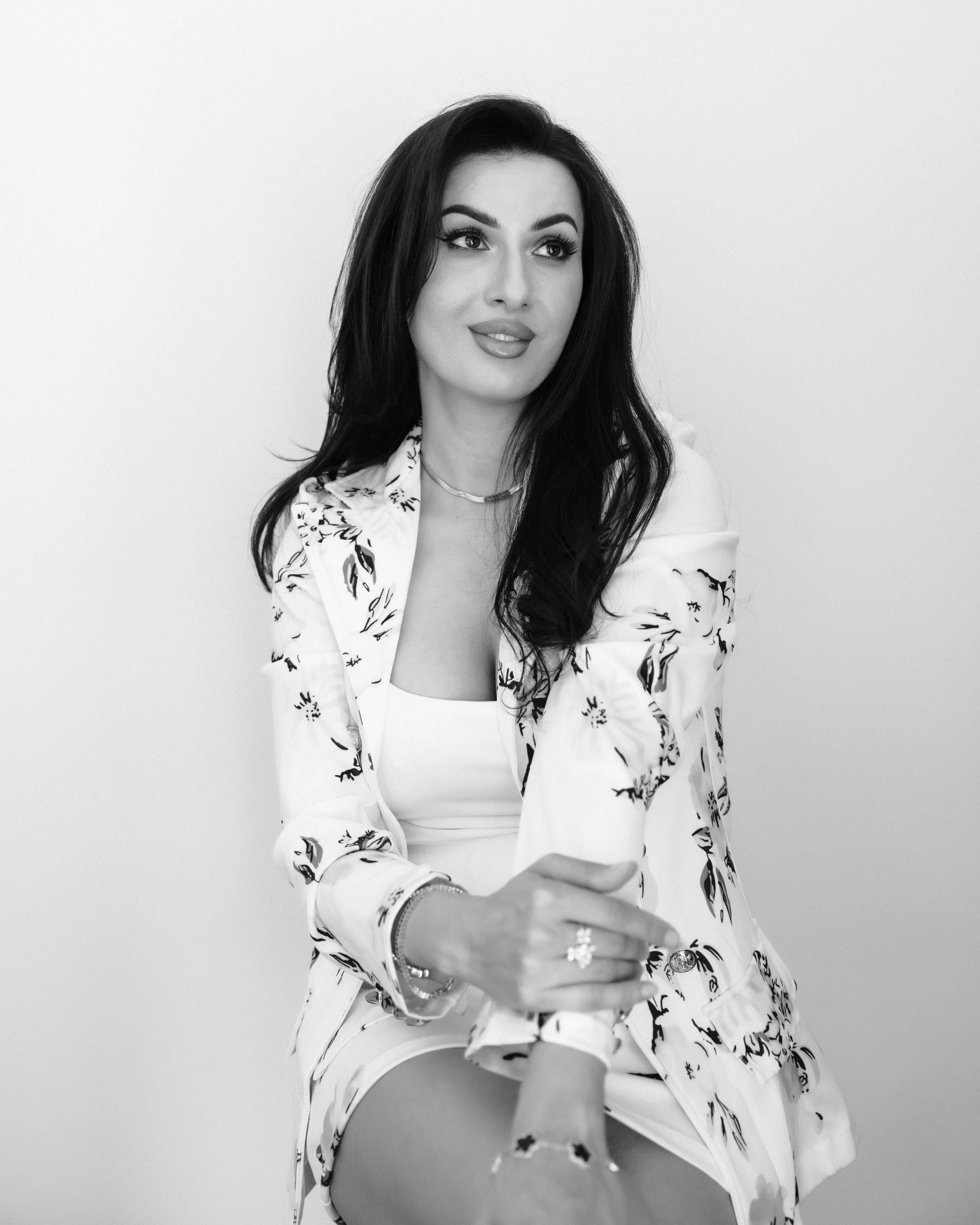 Black-and-white photo of a woman with long dark hair wearing a patterned jacket and jewelry, seated with her hand extended forward, against a plain background.