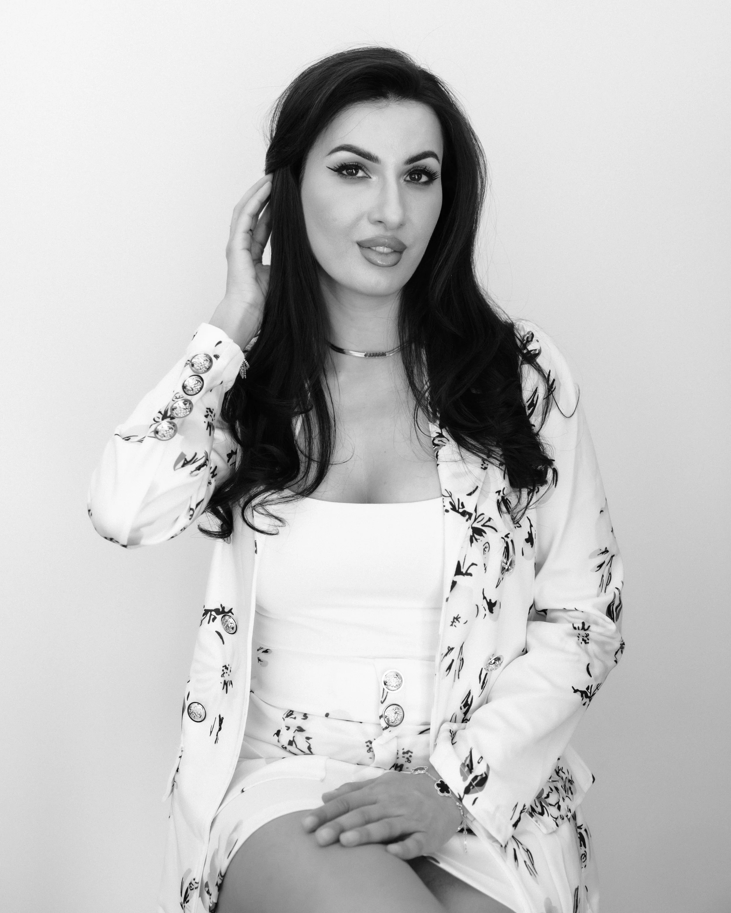 A black-and-white portrait of a woman with long dark hair, wearing a patterned blazer with large buttons, a white top, and jewelry, seated against a plain background.