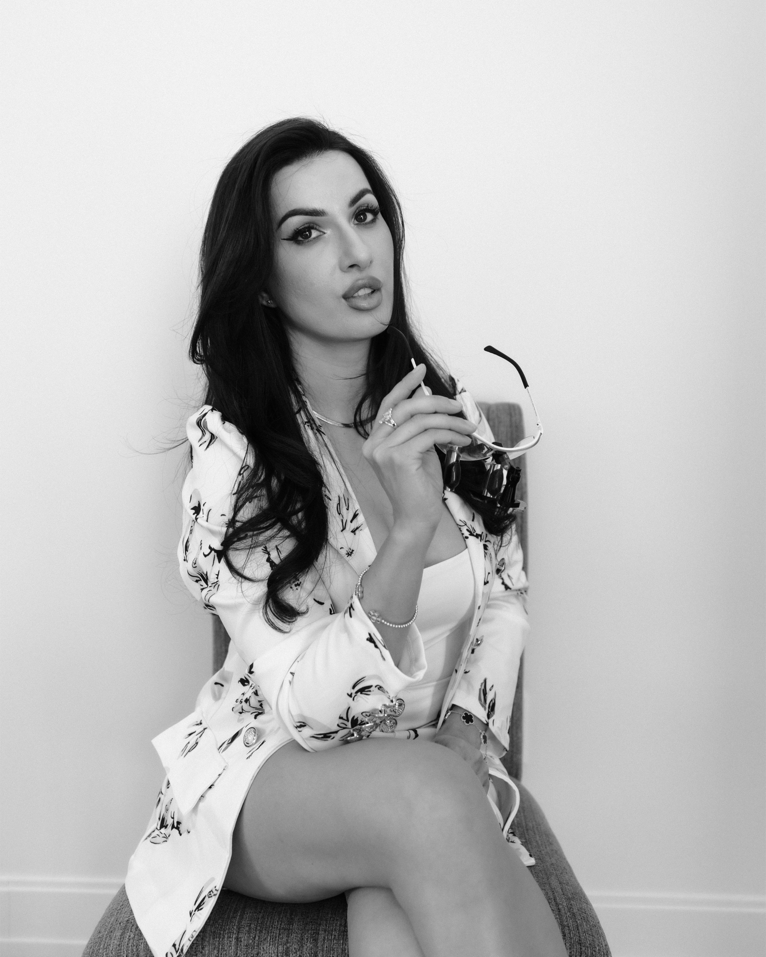 A black and white photo of a woman sitting on a chair, holding her glasses, looking at the camera with a serious expression. She has long dark hair, styled makeup, and is wearing a patterned blazer over a top with her legs crossed.