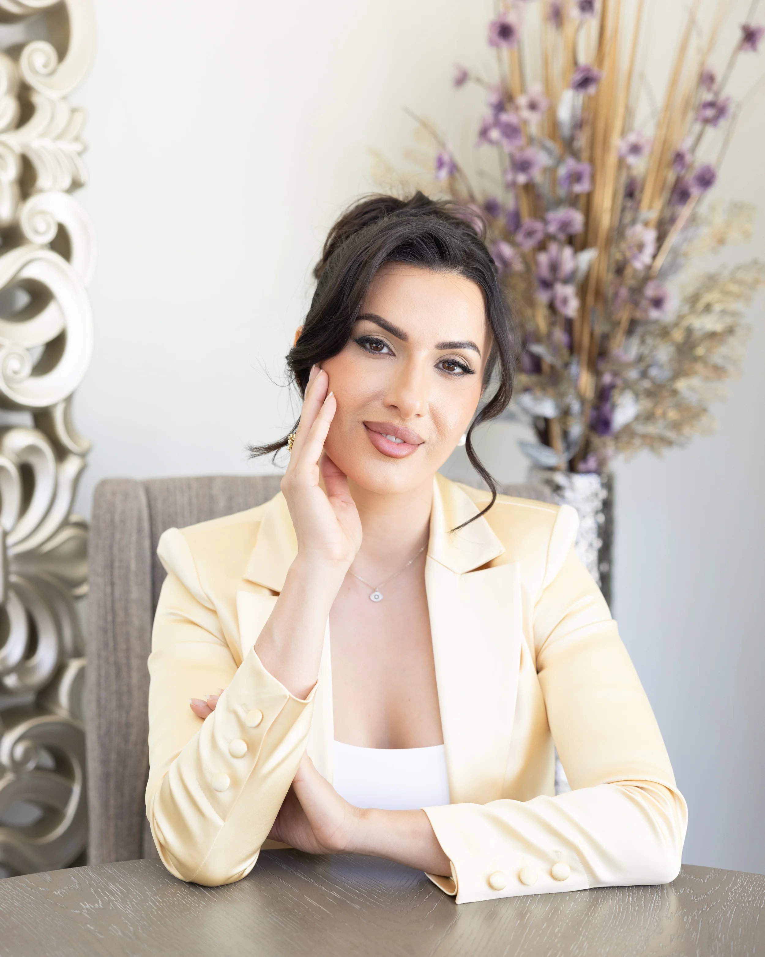 A woman with dark hair and makeup wearing a light yellow blazer, sitting at a wooden table with her hand resting on her cheek, in front of a blurred background with purple flowers and decorative elements.