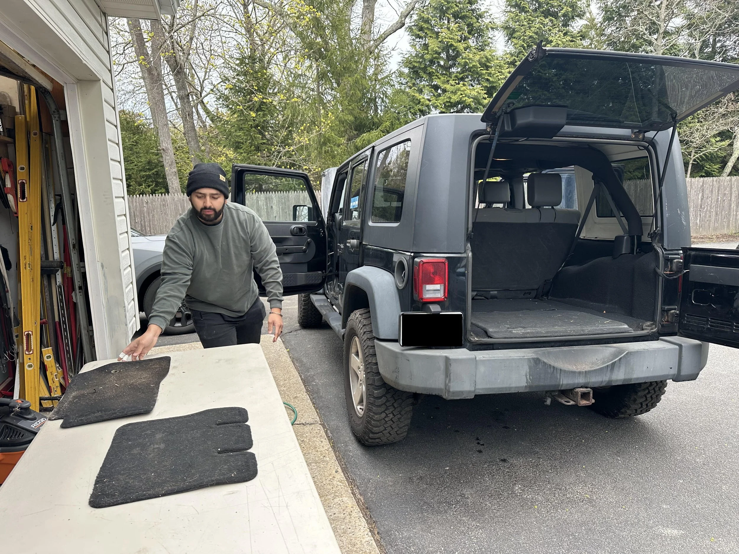 A man in a black beanie and gray sweatshirt is working outside near a silver Jeep with the rear hatch open, next to a white mobile work cabinet with tools and mats on top. Trees and a wooden fence are in the background.