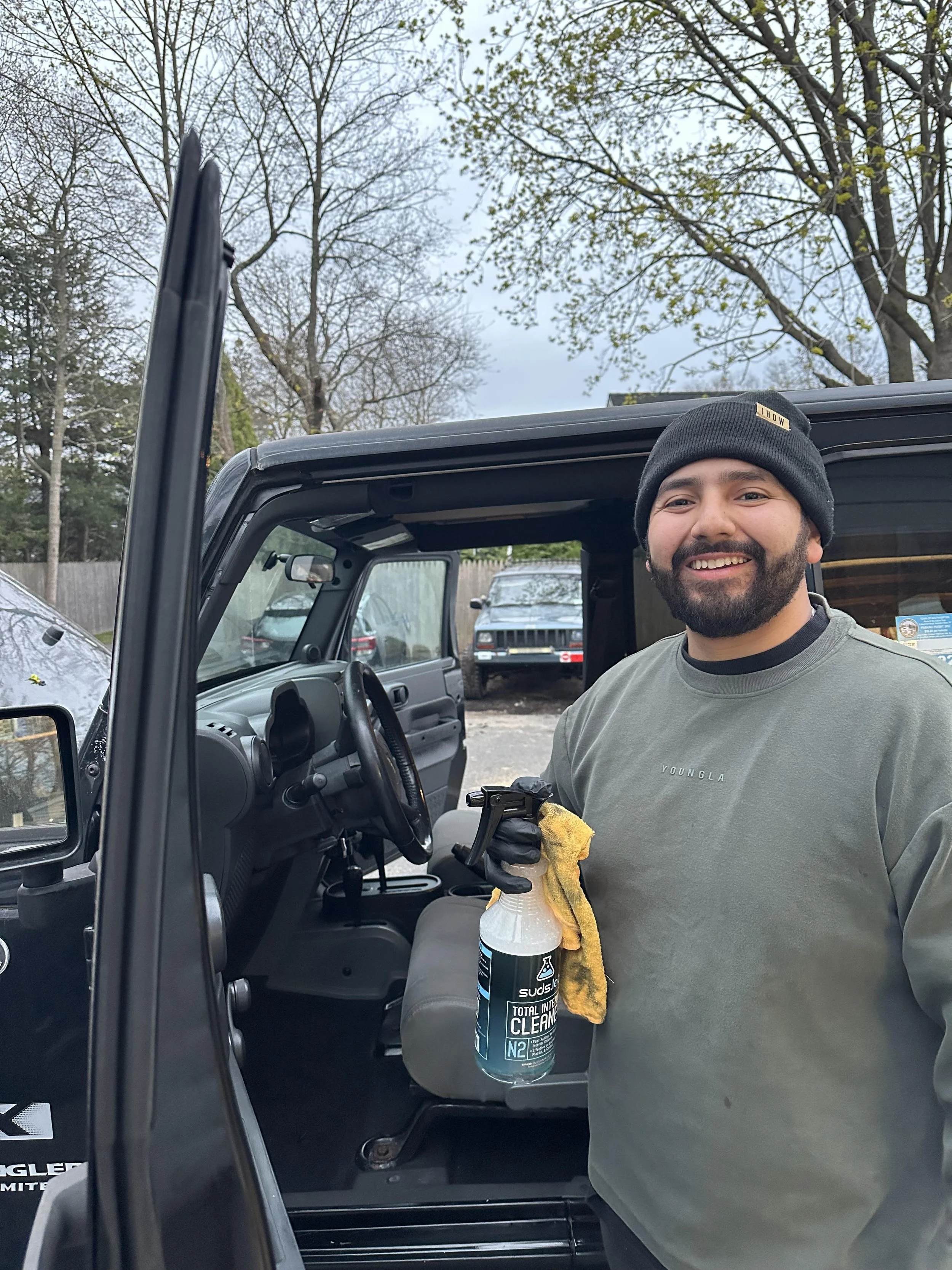Man with beard wearing a black beanie and gray shirt inside a vehicle, holding a cleaning spray and cloth, with trees and parked cars in the background.