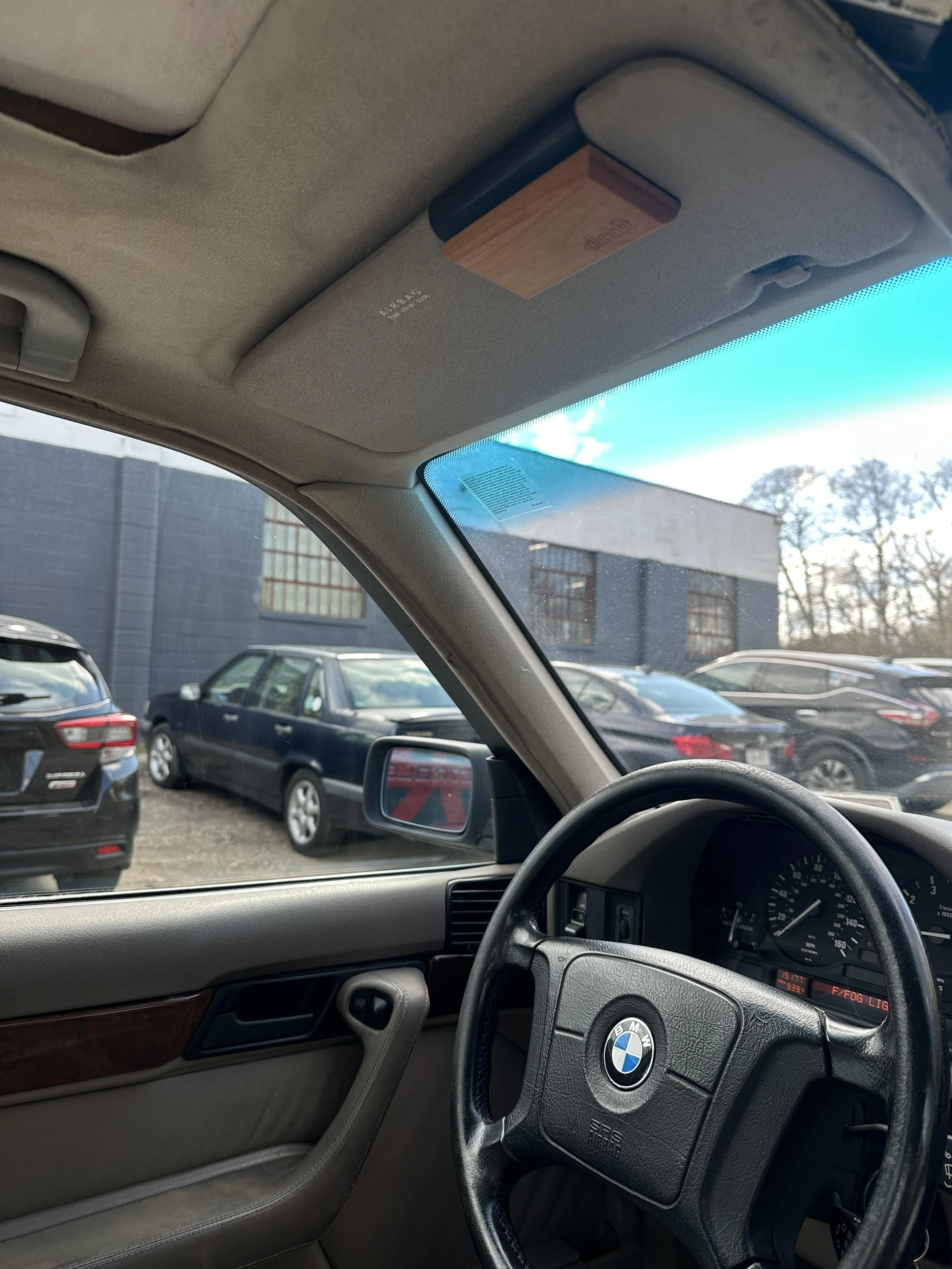 Inside a car viewed from the driver's side, showing the steering wheel with a BMW logo, the dashboard, and part of the front passenger seat. Outside the window, several parked cars and a building are visible.