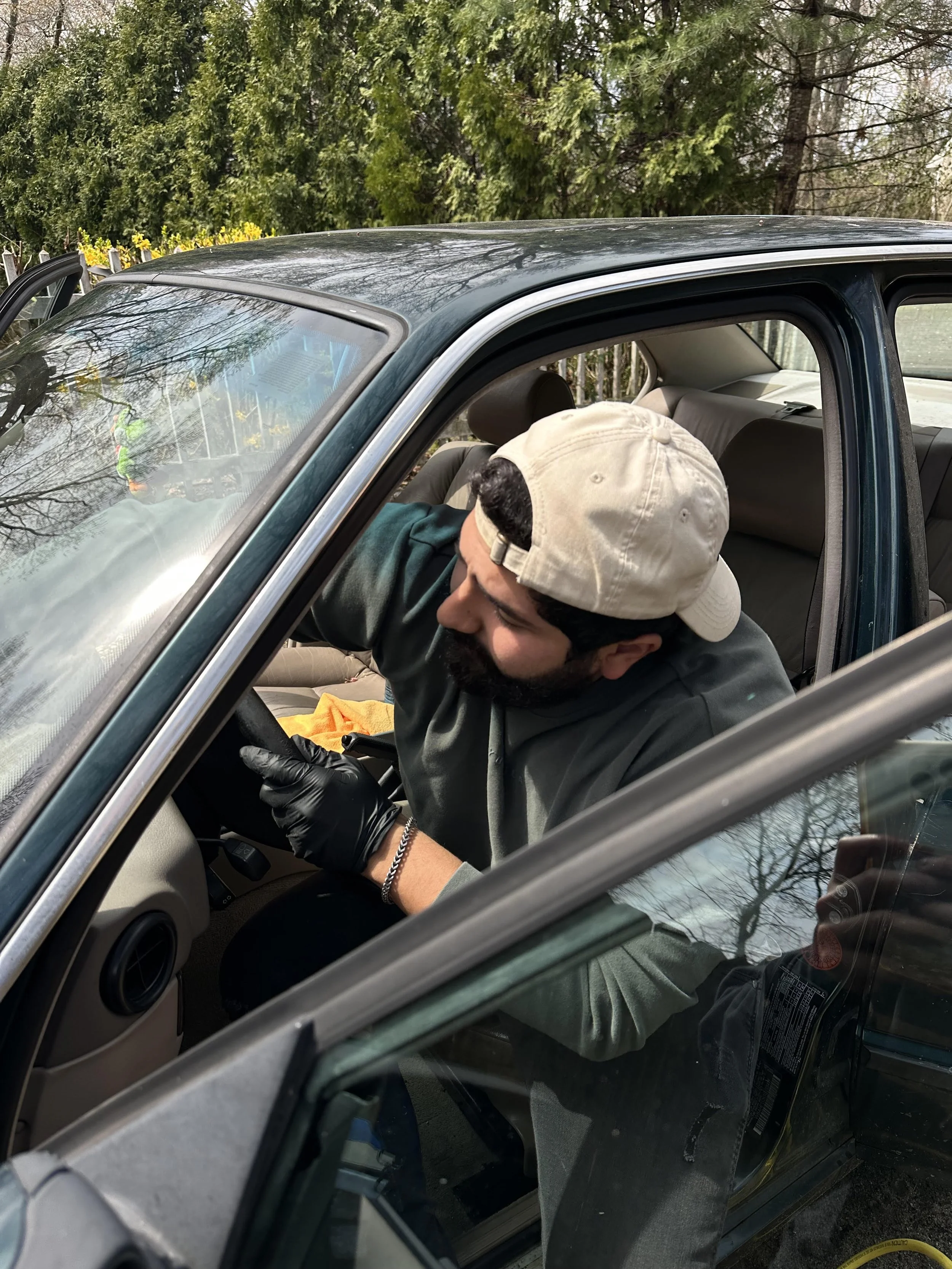 A man wearing a white cap and black gloves leaning into the driver side of a dark green car, with the window down. He appears to be cleaning or inspecting the interior of the vehicle.