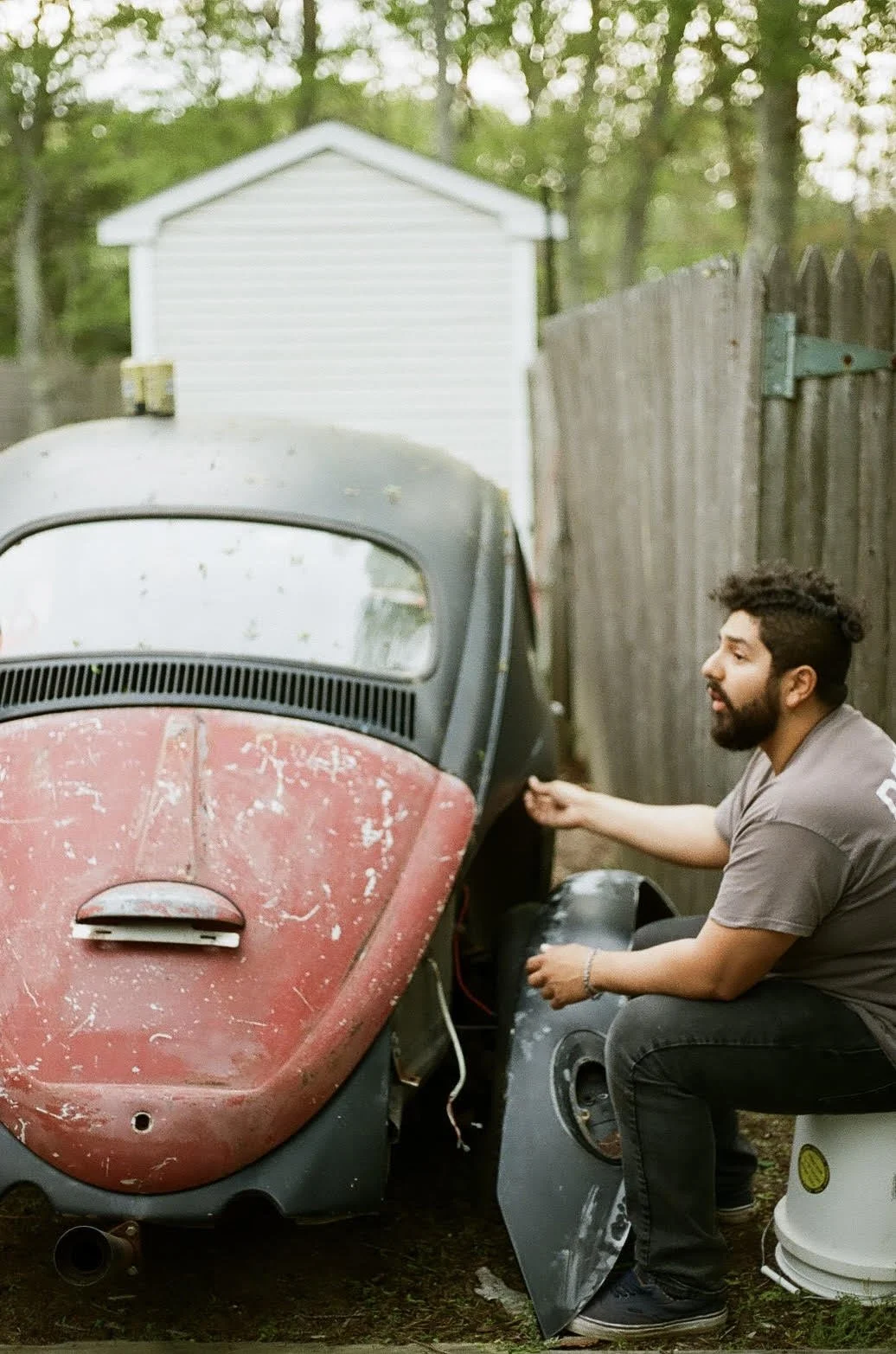 A man with a beard and curly hair sitting on a white bucket in a backyard, working on repairing or restoring a vintage Volkswagen Beetle car with a red hood that is weathered and scratched.