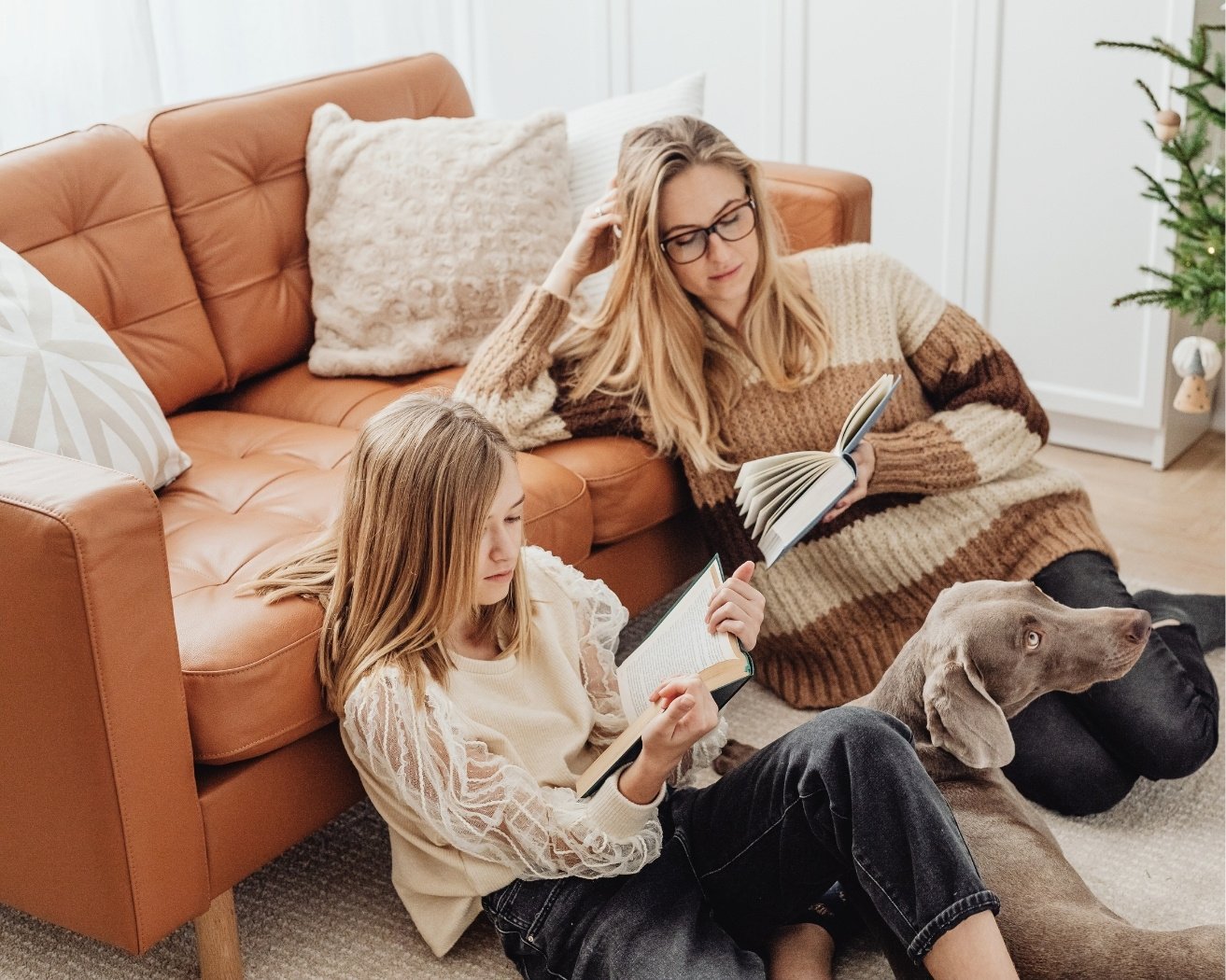 A woman and a girl sitting on the floor and reading books, with a dog lying beside them, in a cozy living room decorated for Christmas.