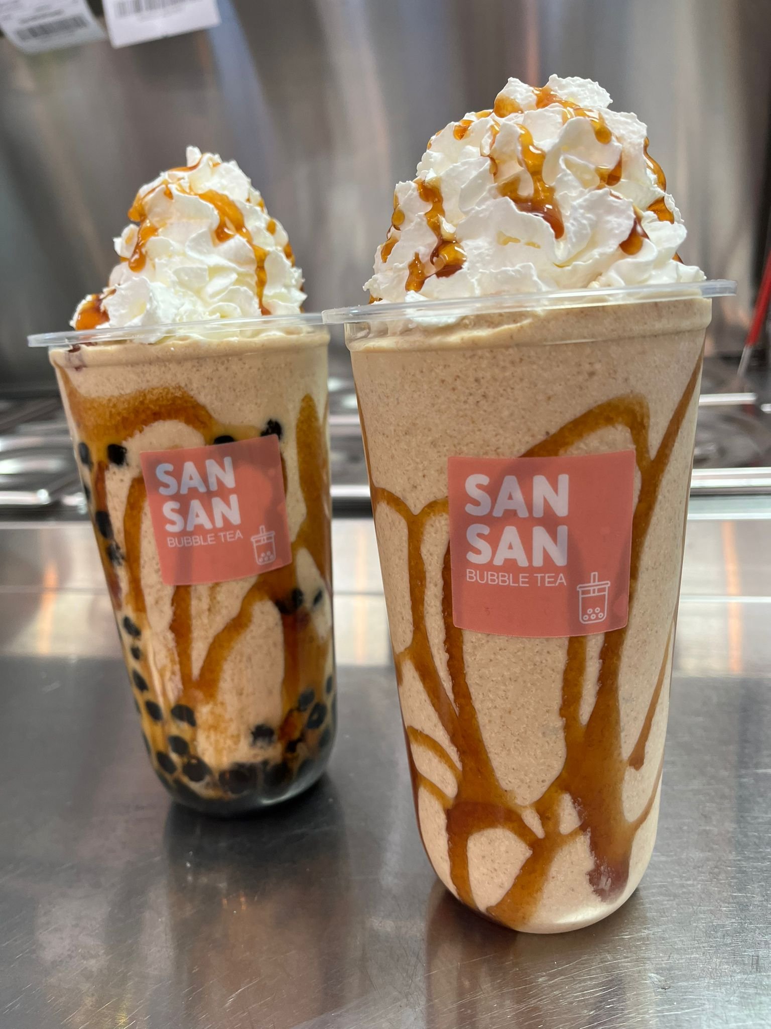 Two bubble tea drinks with whipped cream and caramel drizzle, one with tapioca pearls at the bottom, on a metallic surface.