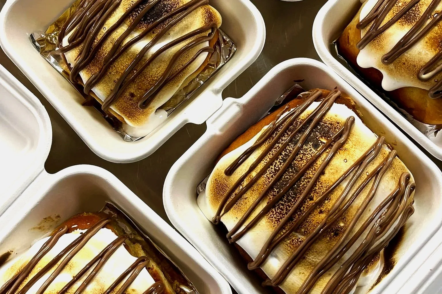 Four containers holding baked s'mores bars topped with chocolate drizzle.