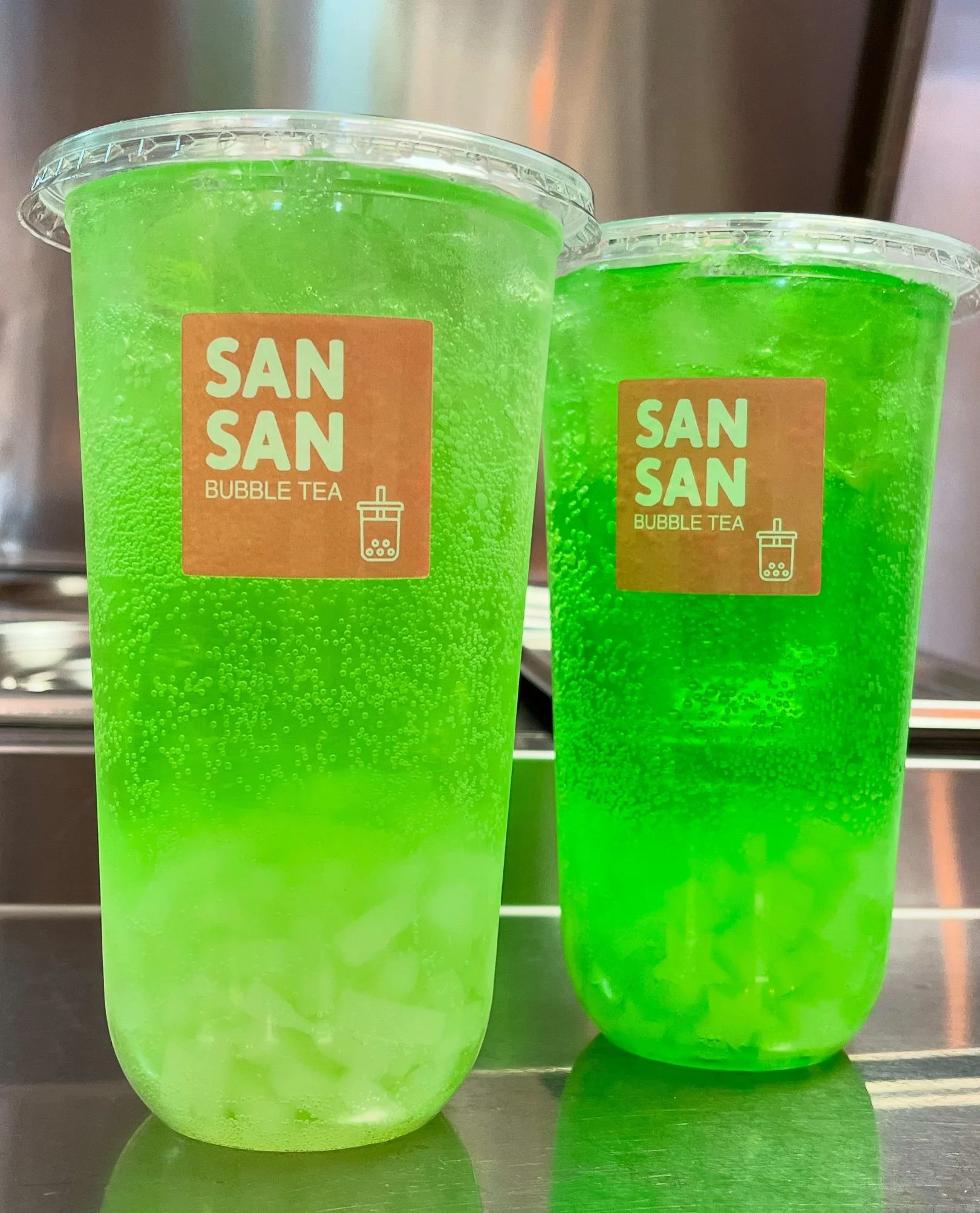 Two tall, clear cups filled with bright green bubble tea, labeled 'San San Bubble Tea,' with a straw illustration, placed on a metallic surface.