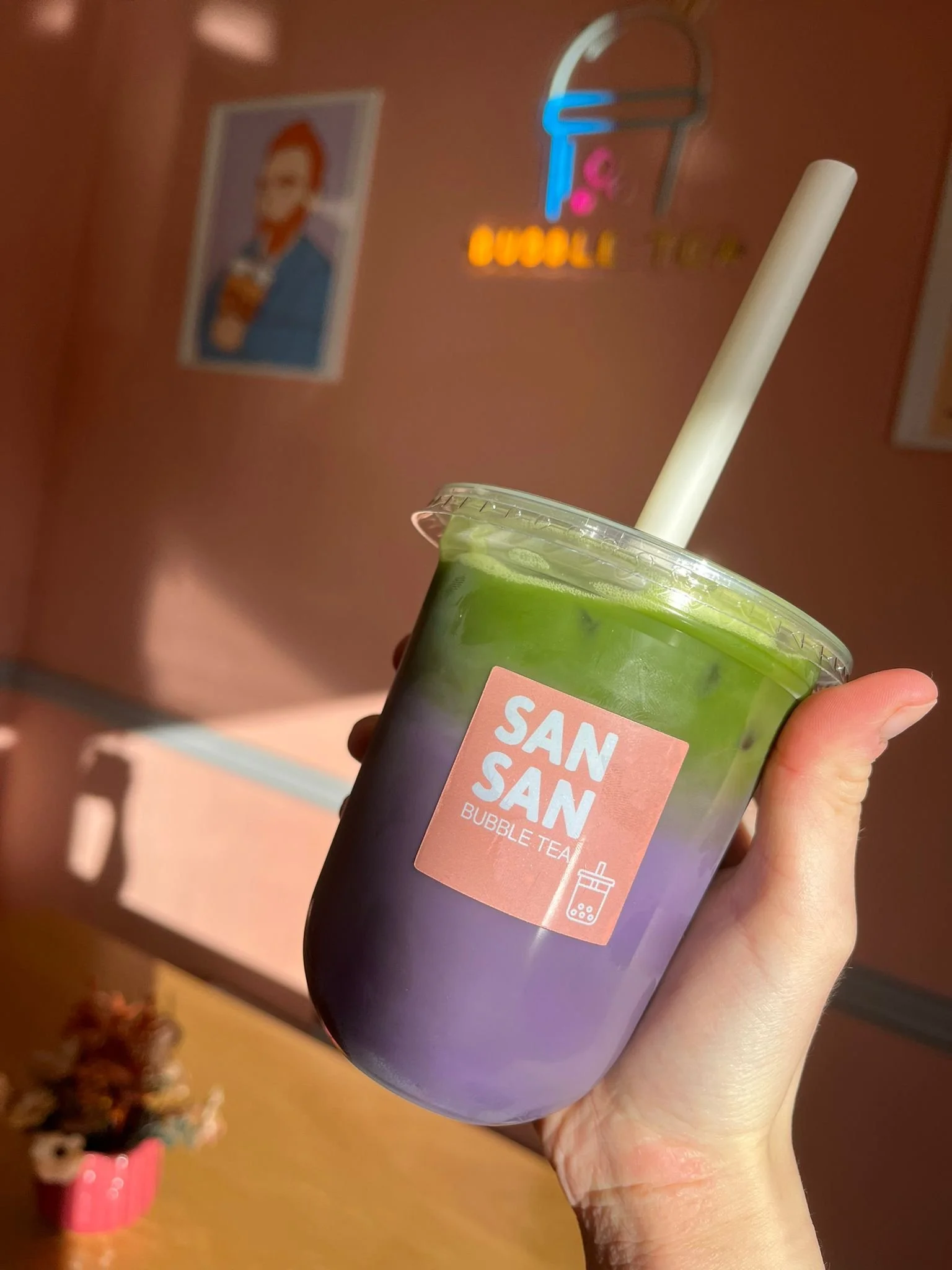 A hand holding a cup of tie-dye bubble tea with layers of purple and green, a straw, in front of a pink wall with a Neon ice cream cone sign and some wall art.