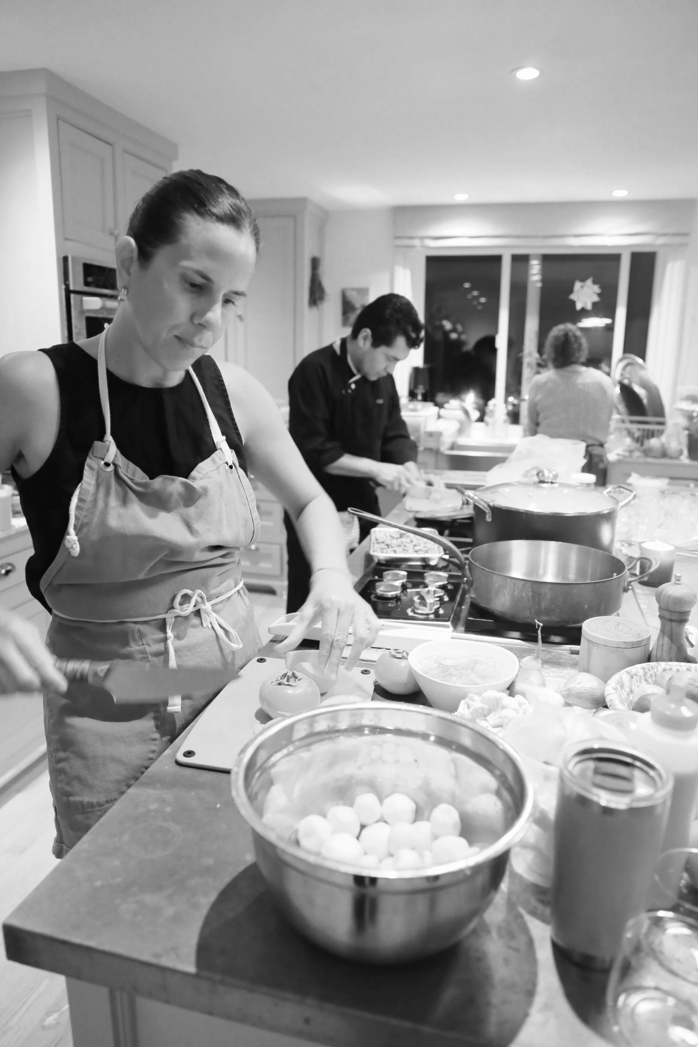 People cooking in a kitchen, woman chopping vegetables and men preparing food on the counter, with ingredients and utensils around.