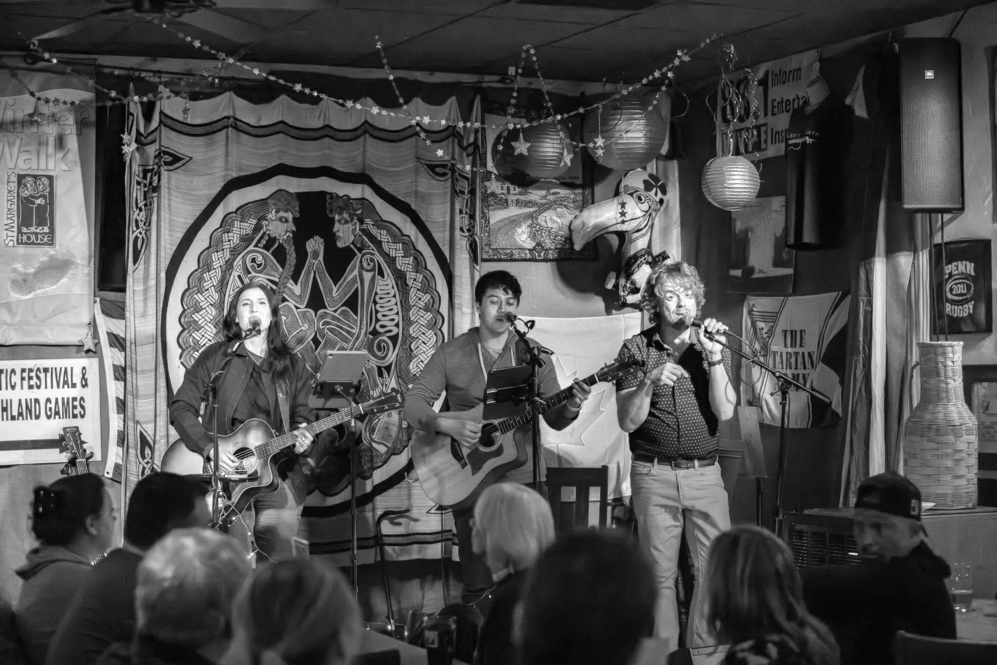 Three musicians performing on a stage inside a bar or music venue. Two women and one man are playing guitars, with the woman on the left and the middle and the man on the right singing into microphones. The background features decorative banners, lan