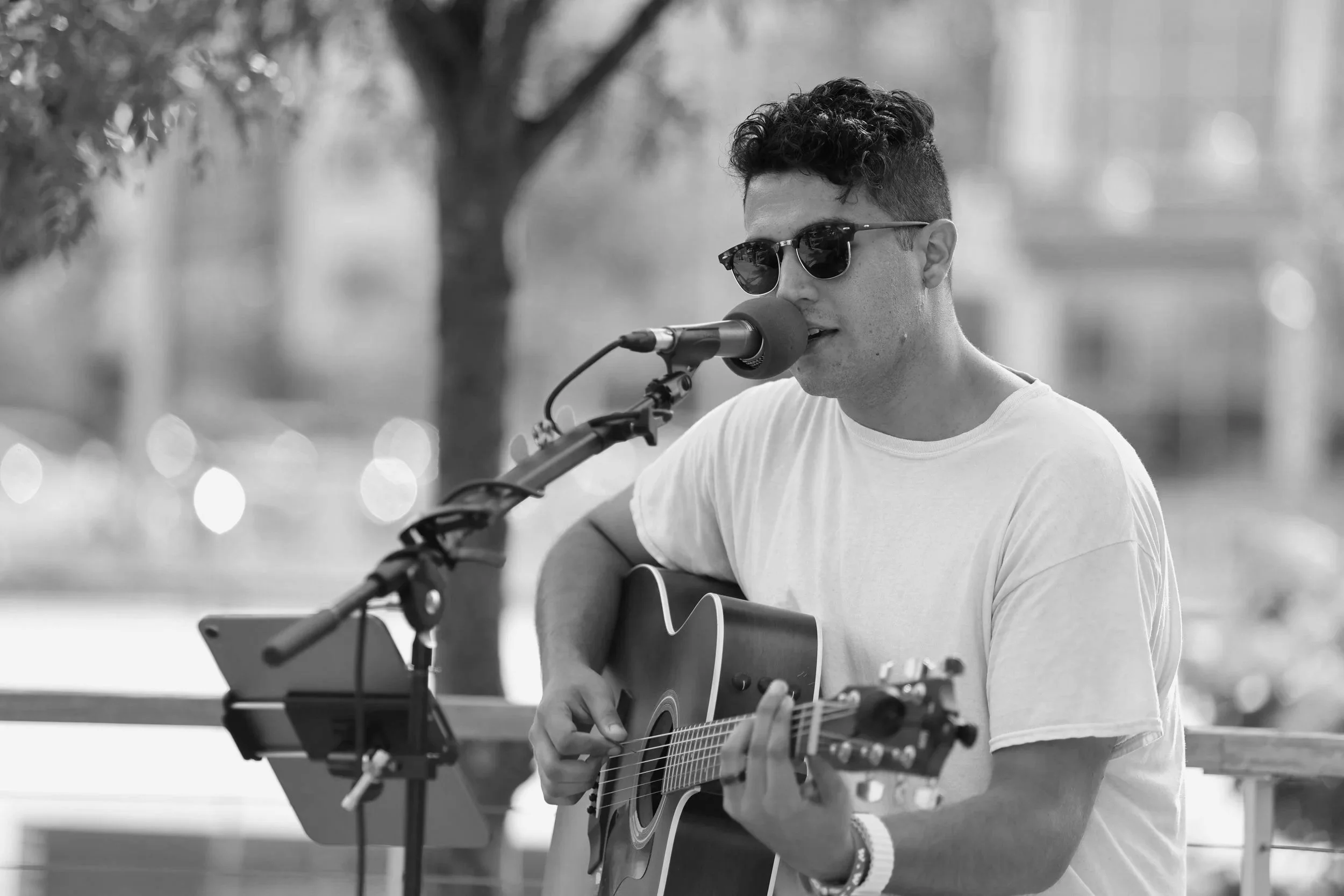A young man with curly hair wearing sunglasses and a white t-shirt, singing into a microphone while playing an acoustic guitar outdoors.