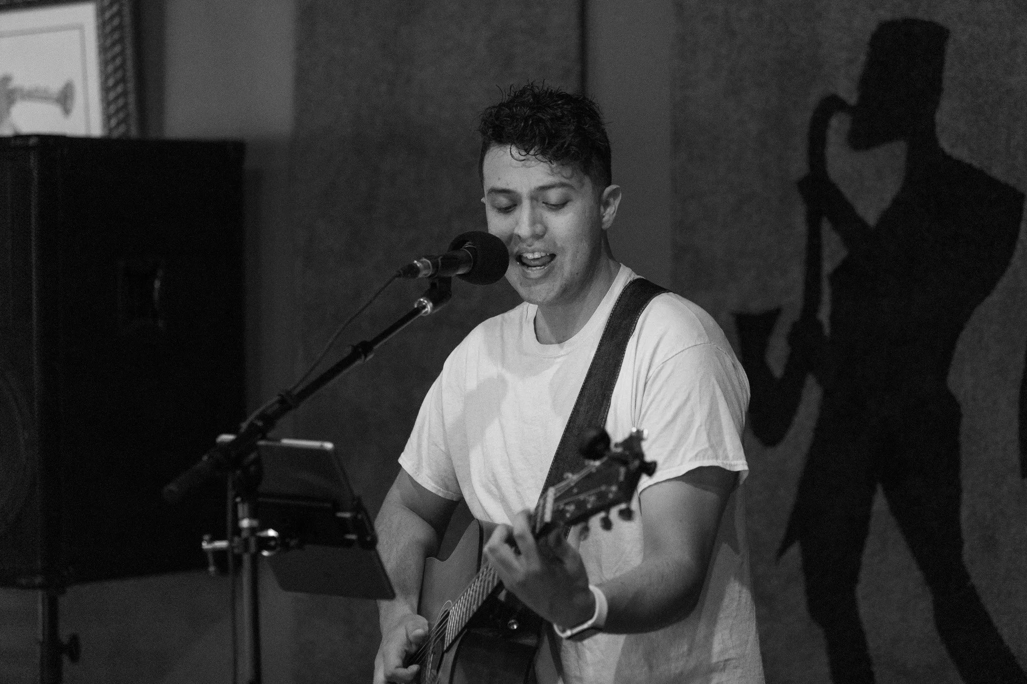 A young man with short, curly hair singing into a microphone and playing an acoustic guitar during a performance in a dimly lit venue. There is a silhouette of a singer with a microphone painted on the wall behind him.