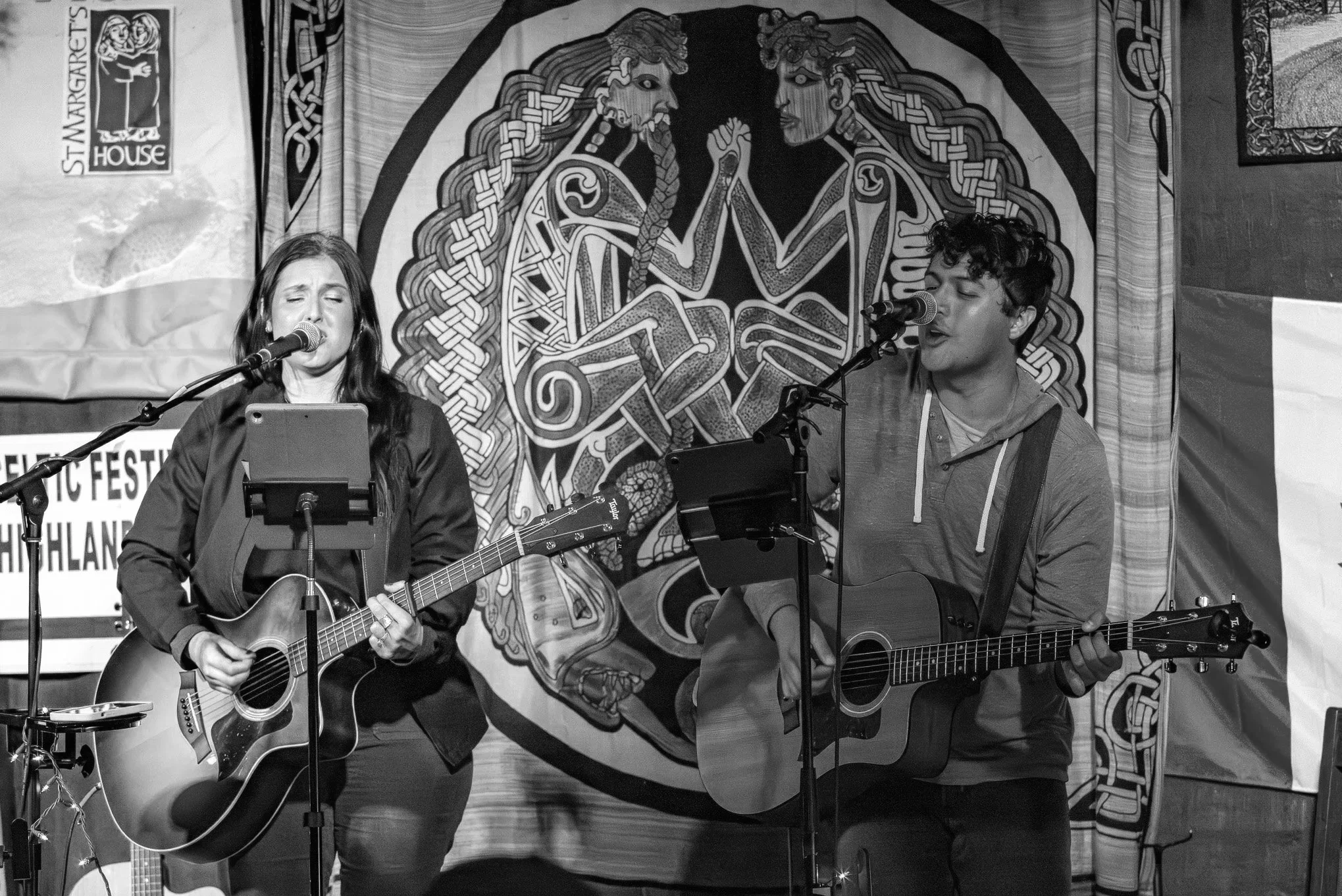 Two musicians performing on stage with acoustic guitars, singing into microphones, in front of a Celtic knot and mythological creature mural at a music festival.