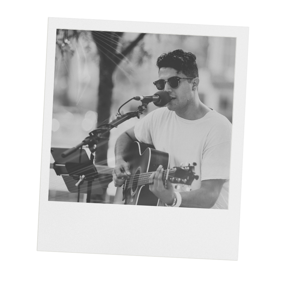 A black and white photograph of a young man wearing sunglasses, playing an acoustic guitar and singing into a microphone outdoors.