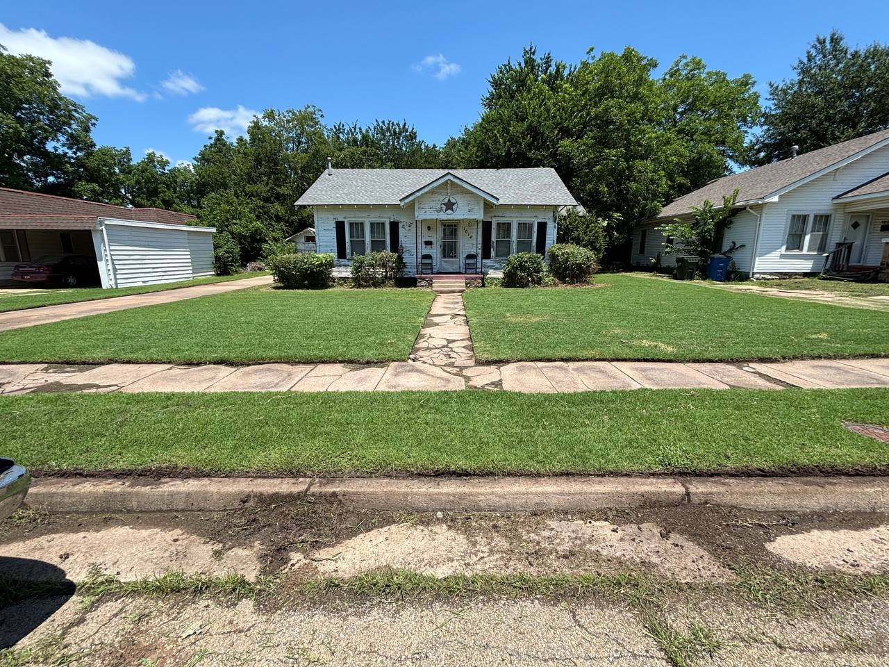 Front yard of a white, weathered house with a small porch, surrounded by green bushes and grass, under a clear blue sky with a few clouds.