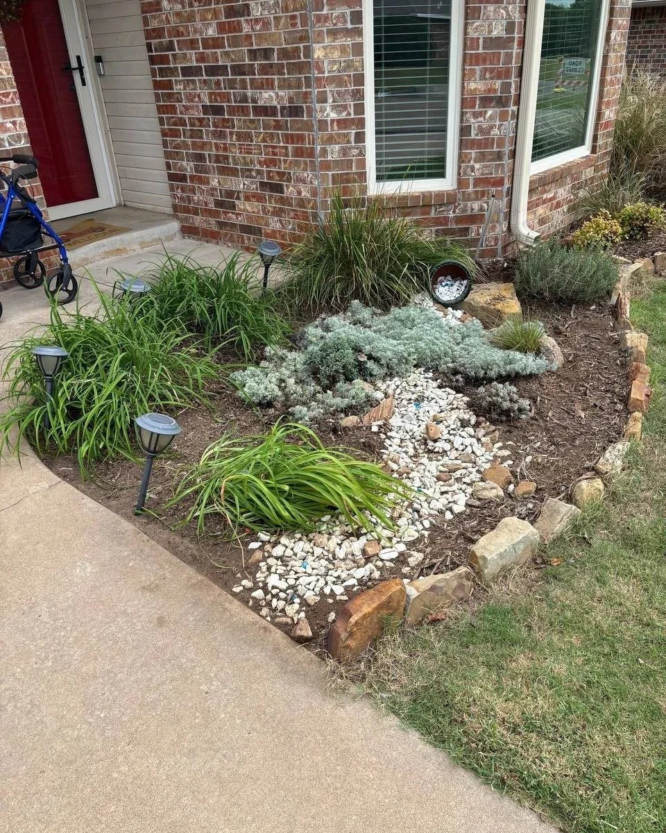 Front yard garden with various plants, white rocks, small solar lights, and a brick house with windows.