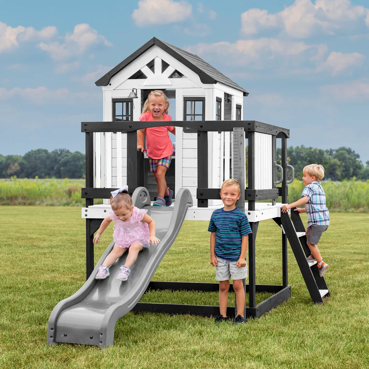 Backyard Discovery Sweetwater Heights wooden playhouse playset with slide and ladder where children are playing in a backyard.