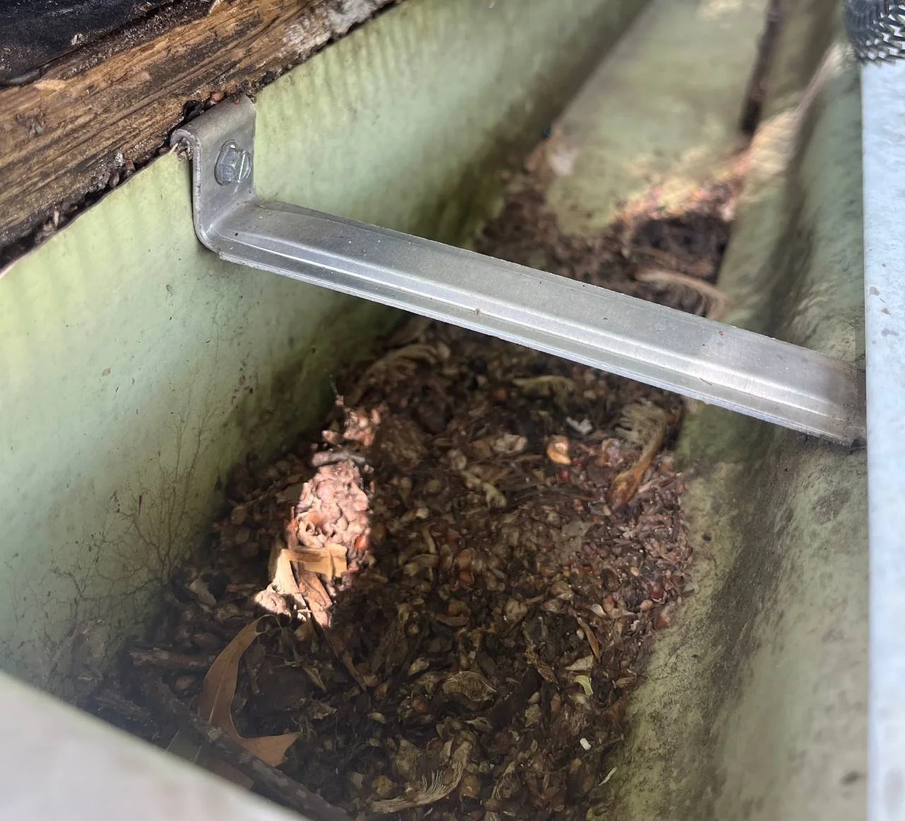 Close-up of a metal cleaning tool inside a weathered green metal container filled with dirt, small stones, and dried leaves, with a wooden top visible at the edge.