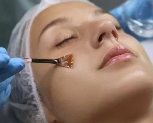 A woman is lying down with her eyes closed, wearing a surgical cap and makeup. A clinician wearing gloves is using a tool on her cheek.