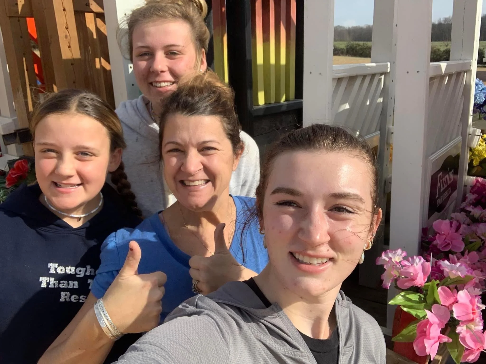 Four women smiling outdoors, taking a selfie at a flower shop, with colorful flowers around them.