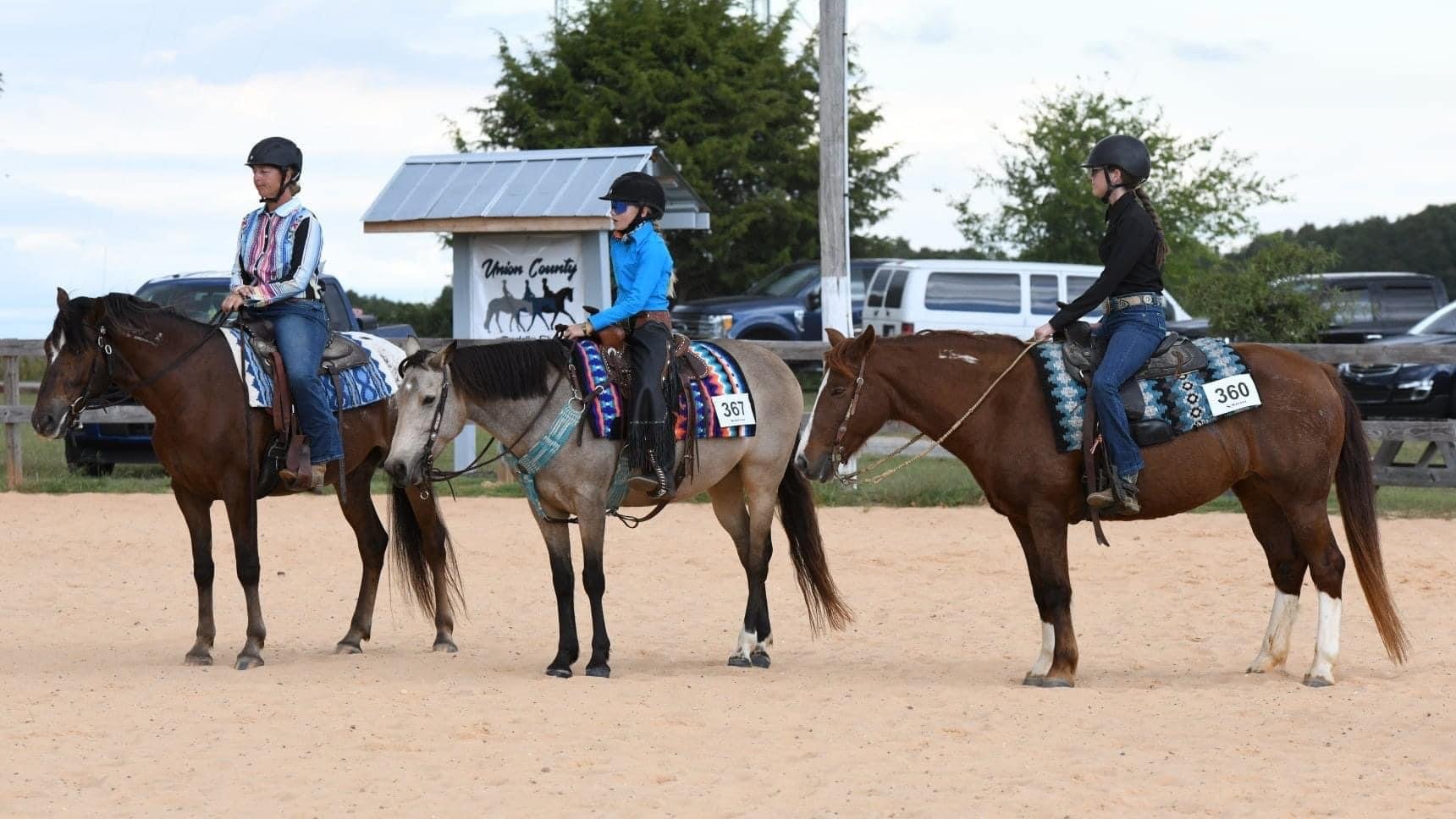 Three women riding horses in an outdoor riding arena, wearing helmets and colorful riding gear. The horses are standing still, and there are cars and a sign with a horse graphic in the background.
