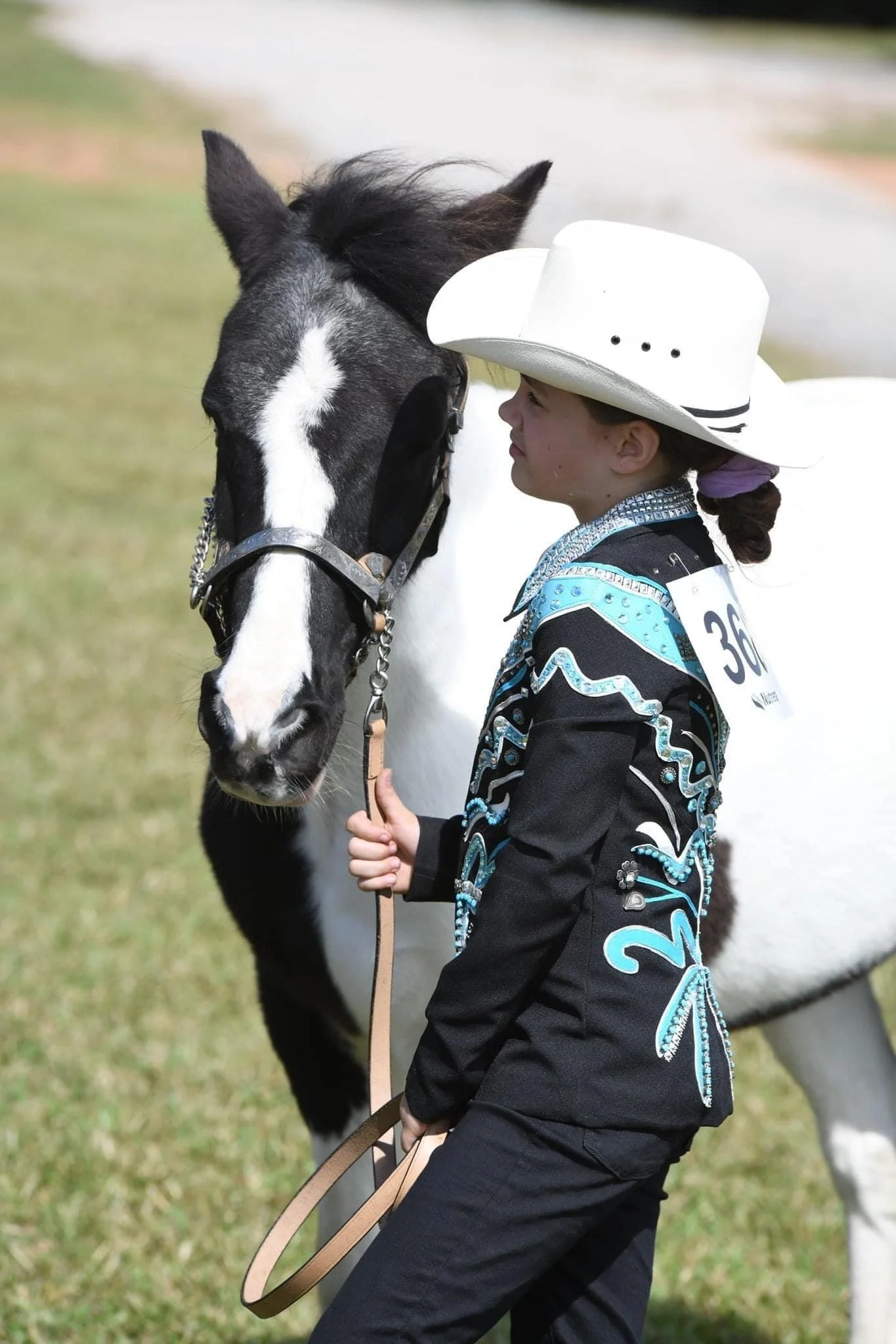 A young girl dressed in western attire, wearing a white cowboy hat, smiling and holding a horse by the reins at an outdoor event.
