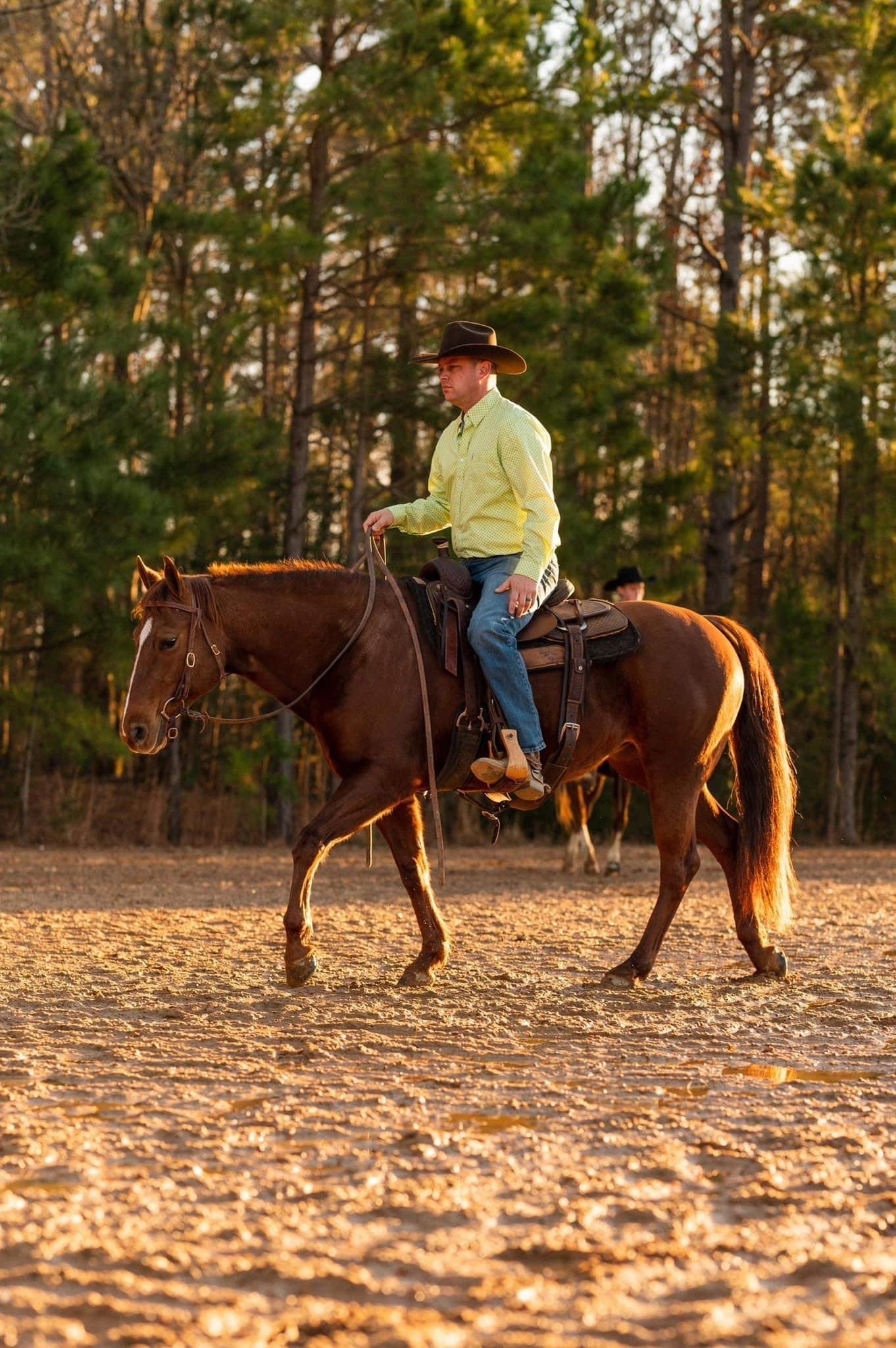 A man wearing a cowboy hat and yellow shirt riding a brown horse on a sandy outdoor field during sunset with trees in the background.