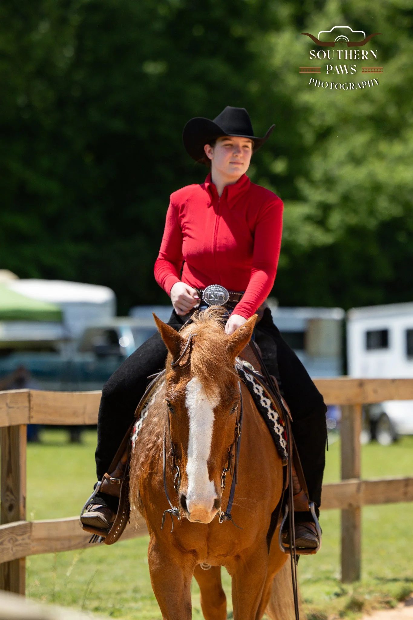 Young woman wearing a red long-sleeve shirt and black cowboy hat riding a chestnut horse with a white blaze, with a wooden fence and green trees in the background.