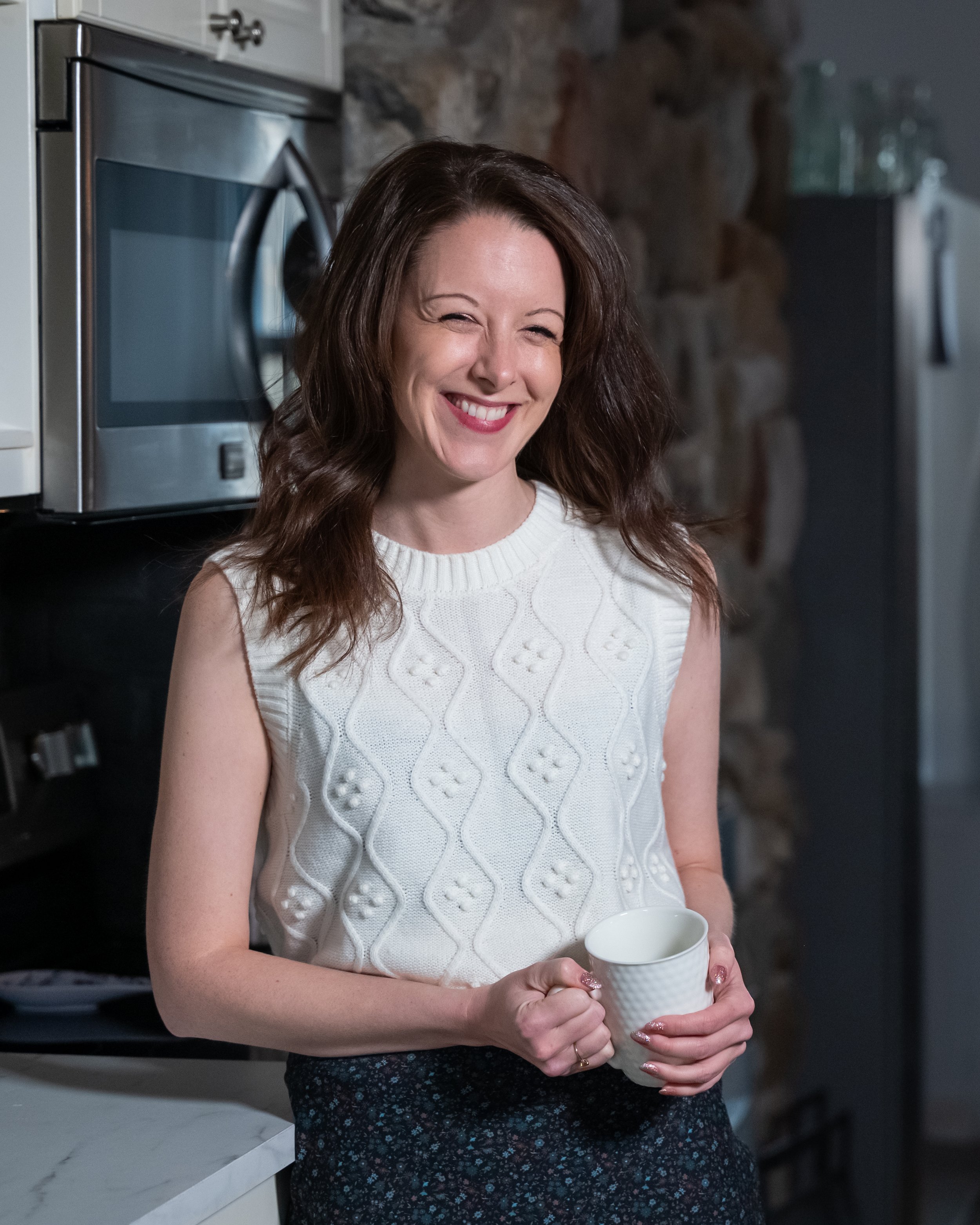 A woman with fair skin, shoulder-length wavy brown hair, wearing a sleeveless white knit sweater with a diamond pattern, is smiling while holding a white mug in a kitchen with a stone wall.