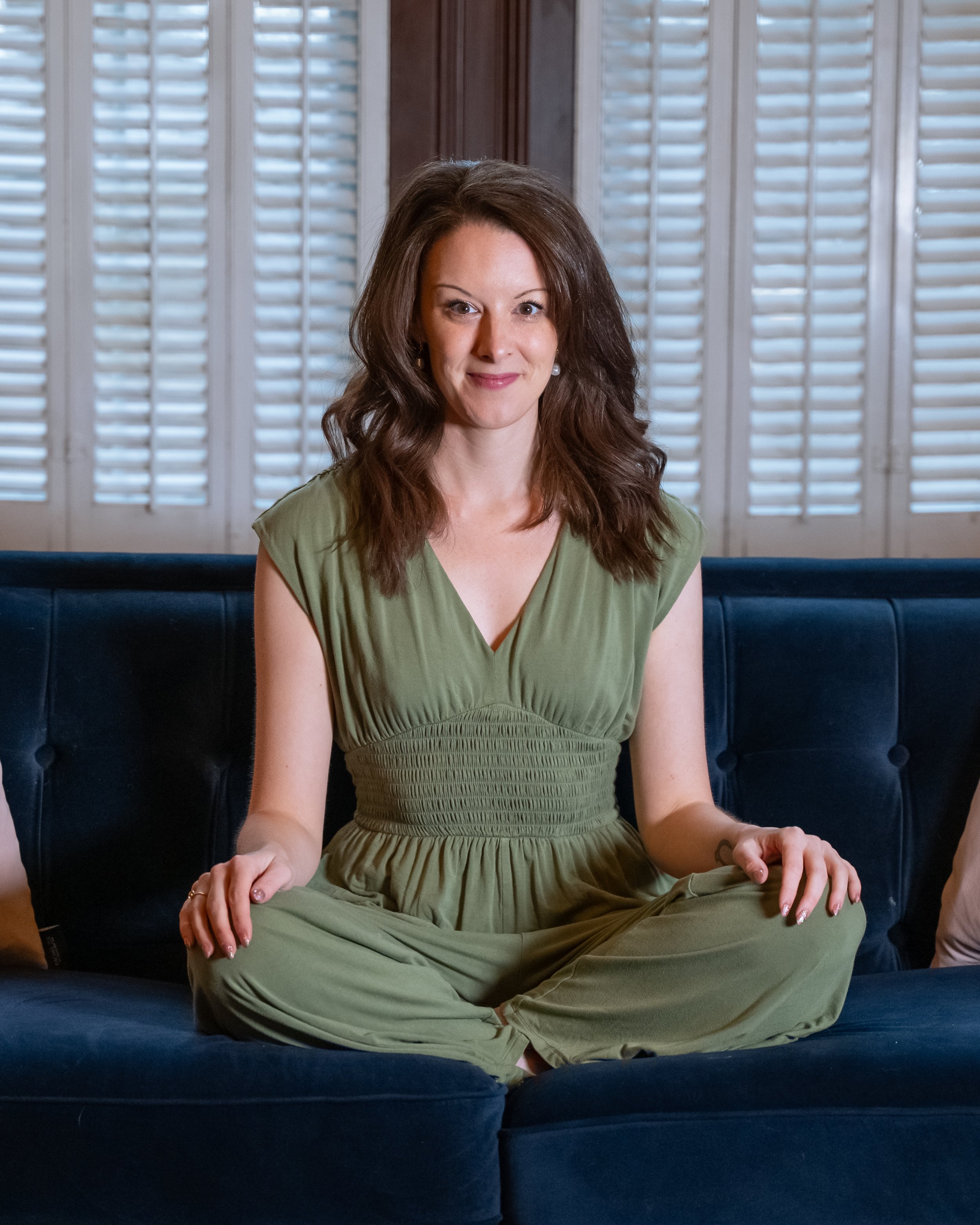 A woman with shoulder-length brown hair sitting cross-legged on a dark blue velvet sofa with white plantation shutters in the background.