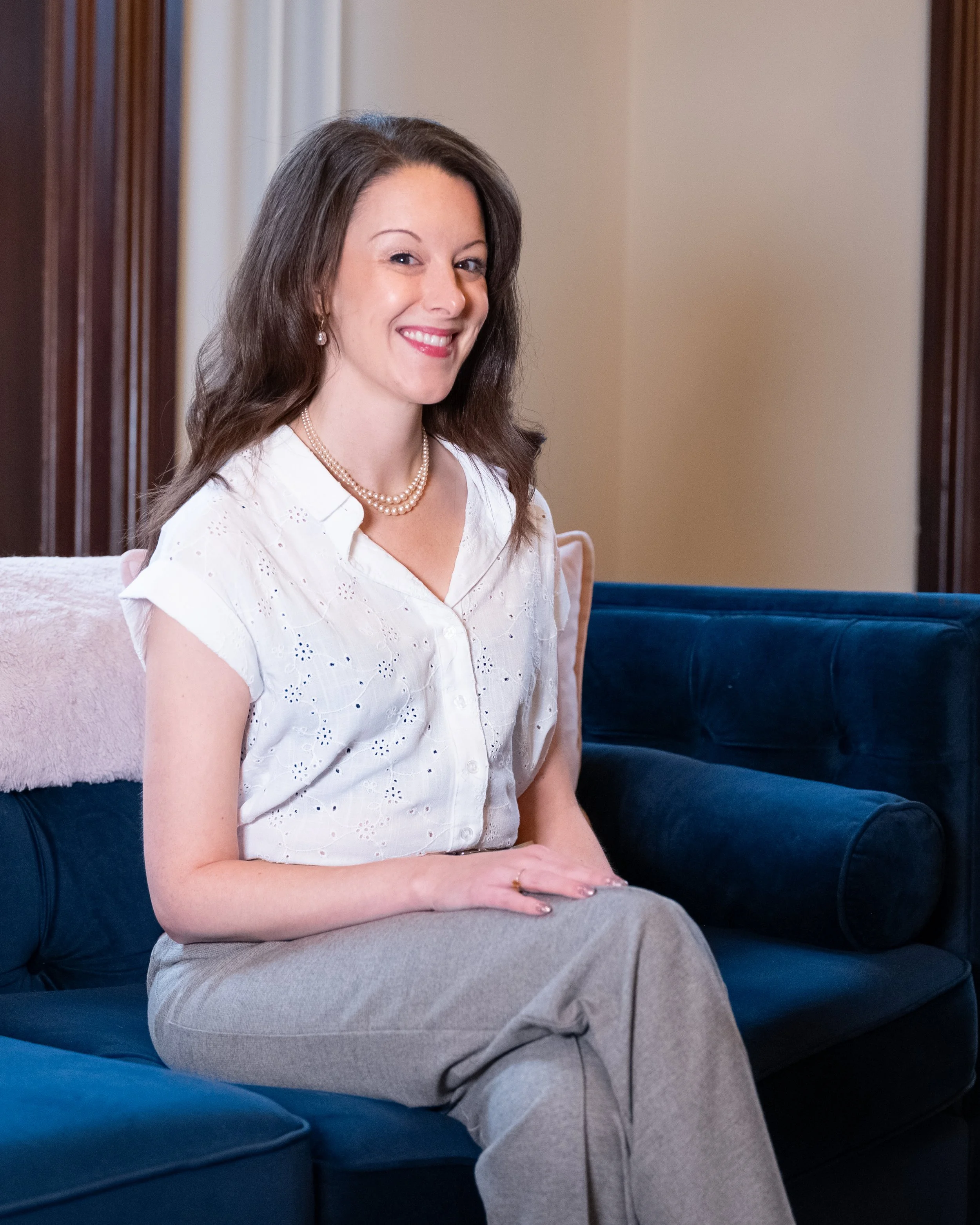 A woman with long dark hair wearing a white blouse, beige pants, pearl necklace and earrings, sitting on a navy blue velvet couch, smiling in a well-lit room with wooden accents.