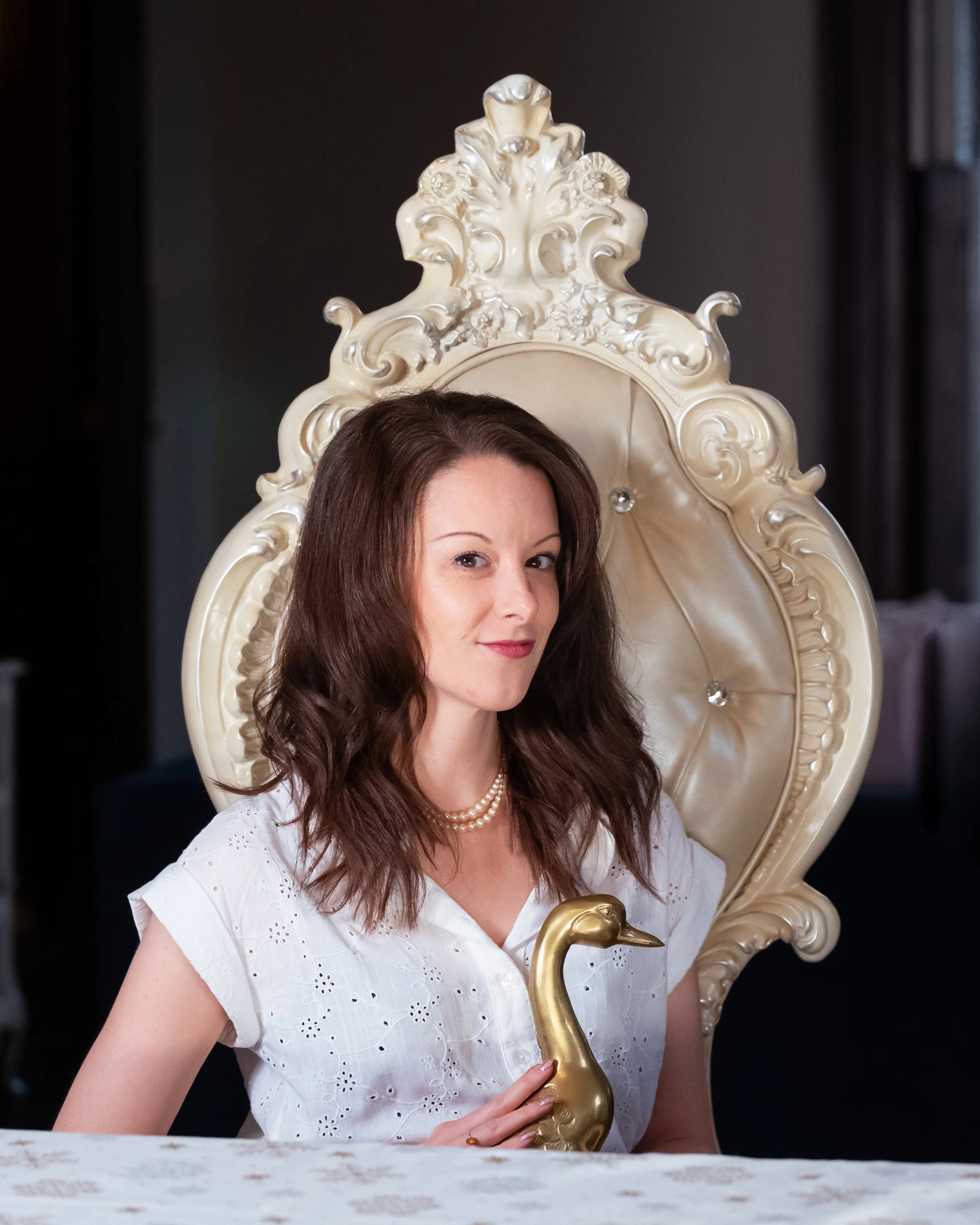 A woman with brown wavy hair in a white dress, sitting on an ornate cream-colored chair with a peacock-shaped handle, holding a golden peacock sculpture.
