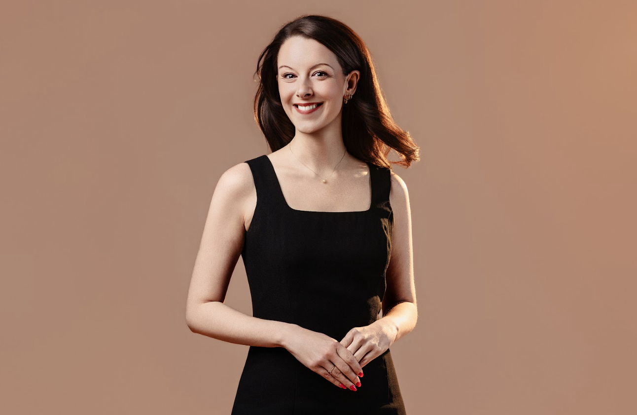 A woman with dark brown hair wearing a black sleeveless dress, standing against a beige background, smiling and looking at the camera.