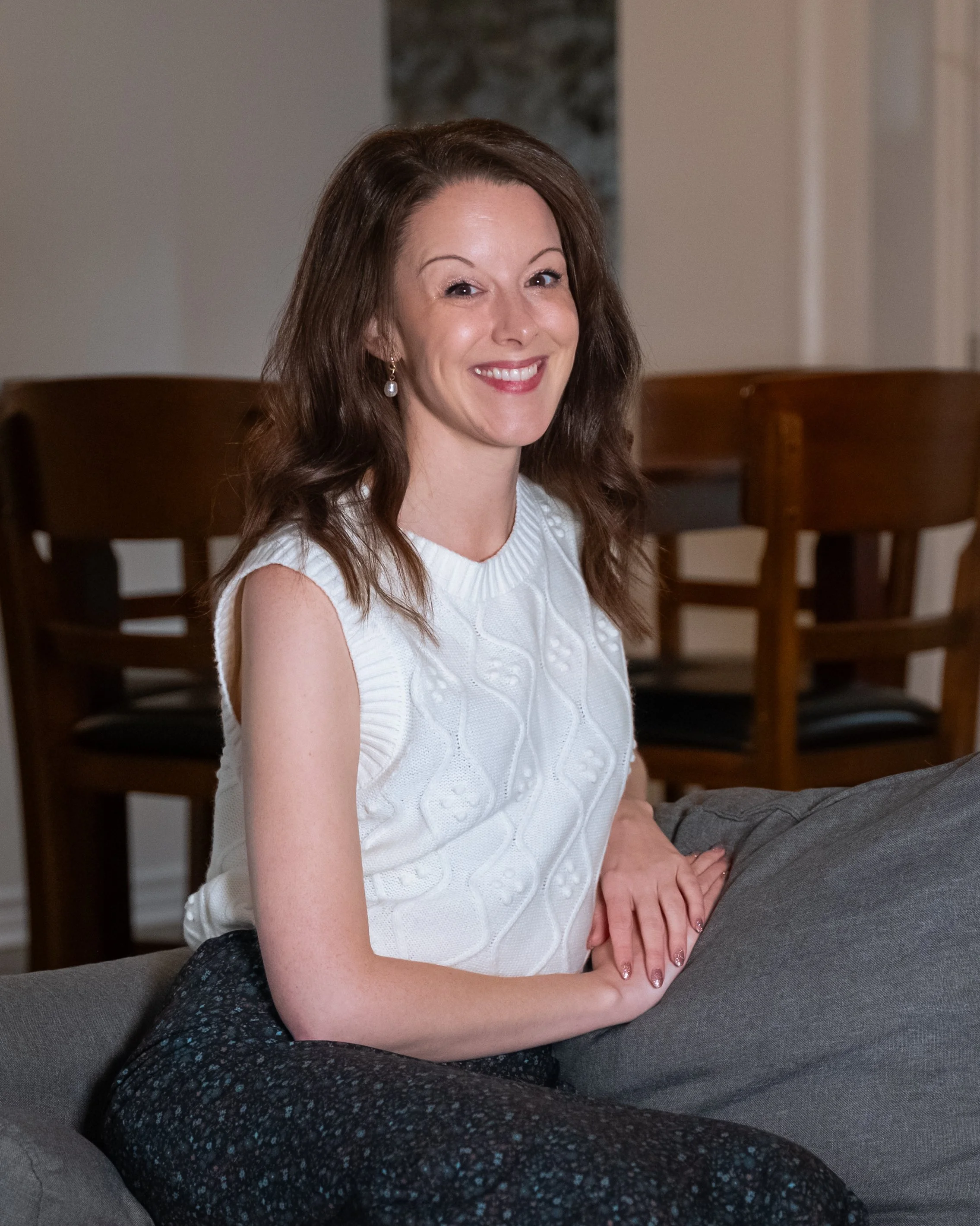 A smiling woman with long brown hair, wearing a white sleeveless sweater and patterned pants, sitting on a grey couch in a living room with wooden chairs in the background.