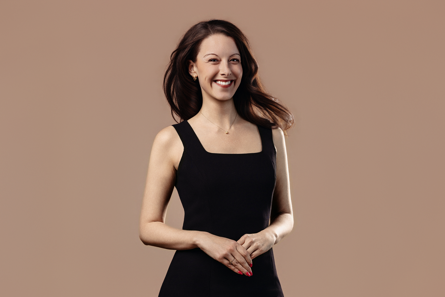 A woman with long brown hair wearing a black sleeveless dress, smiling and standing against a beige background.