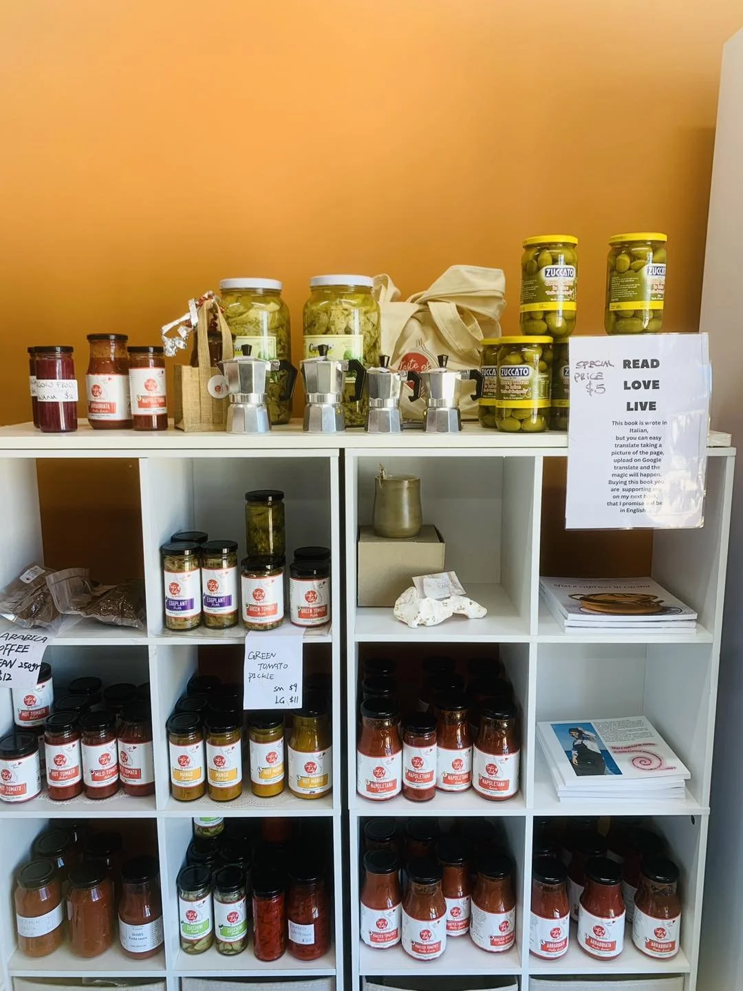 White shelving unit filled with jars of preserved foods and spices, including green olives, tomatoes, and sauces, with a sign displaying prices and a small sign with a quote on the right side.