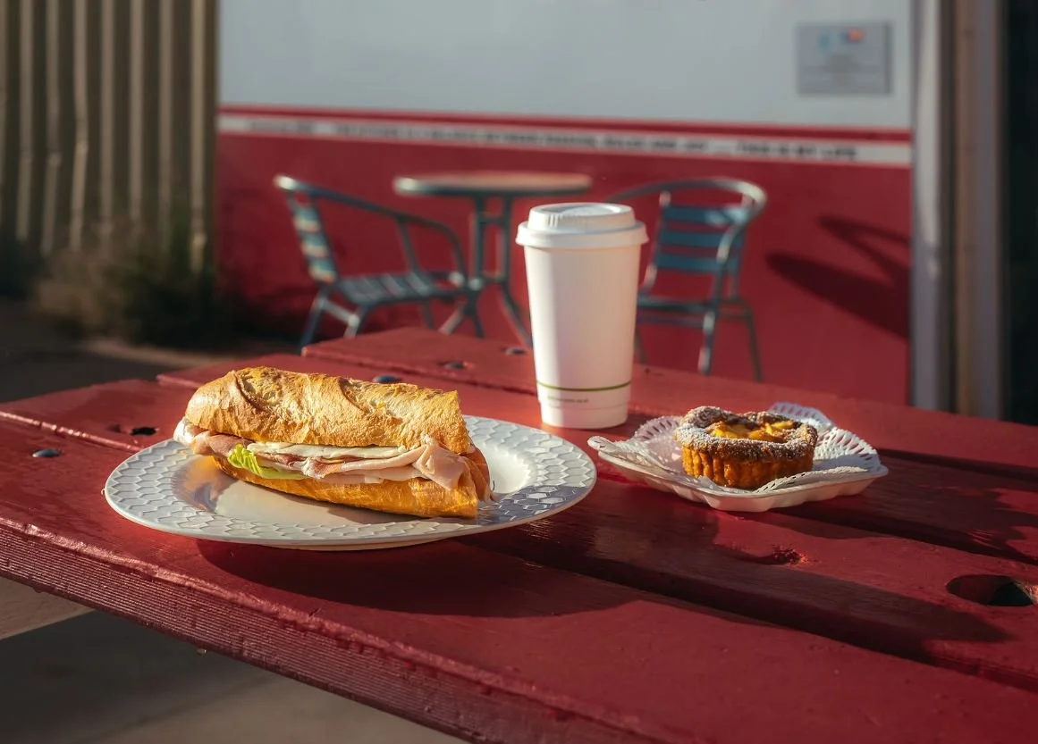 A turkey sandwich on a white paper plate, a muffin on a small paper tray, and a white disposable cup on a red outdoor table.
