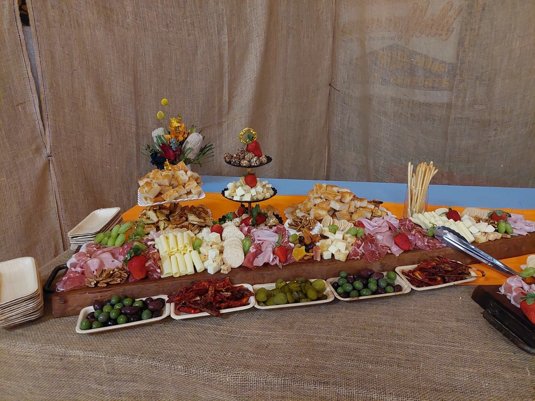 Charcuterie board with cheese, grapes, meats, crackers, and strawberries, decorated with flowers, on a table with paper napkins, in front of a beige fabric backdrop.