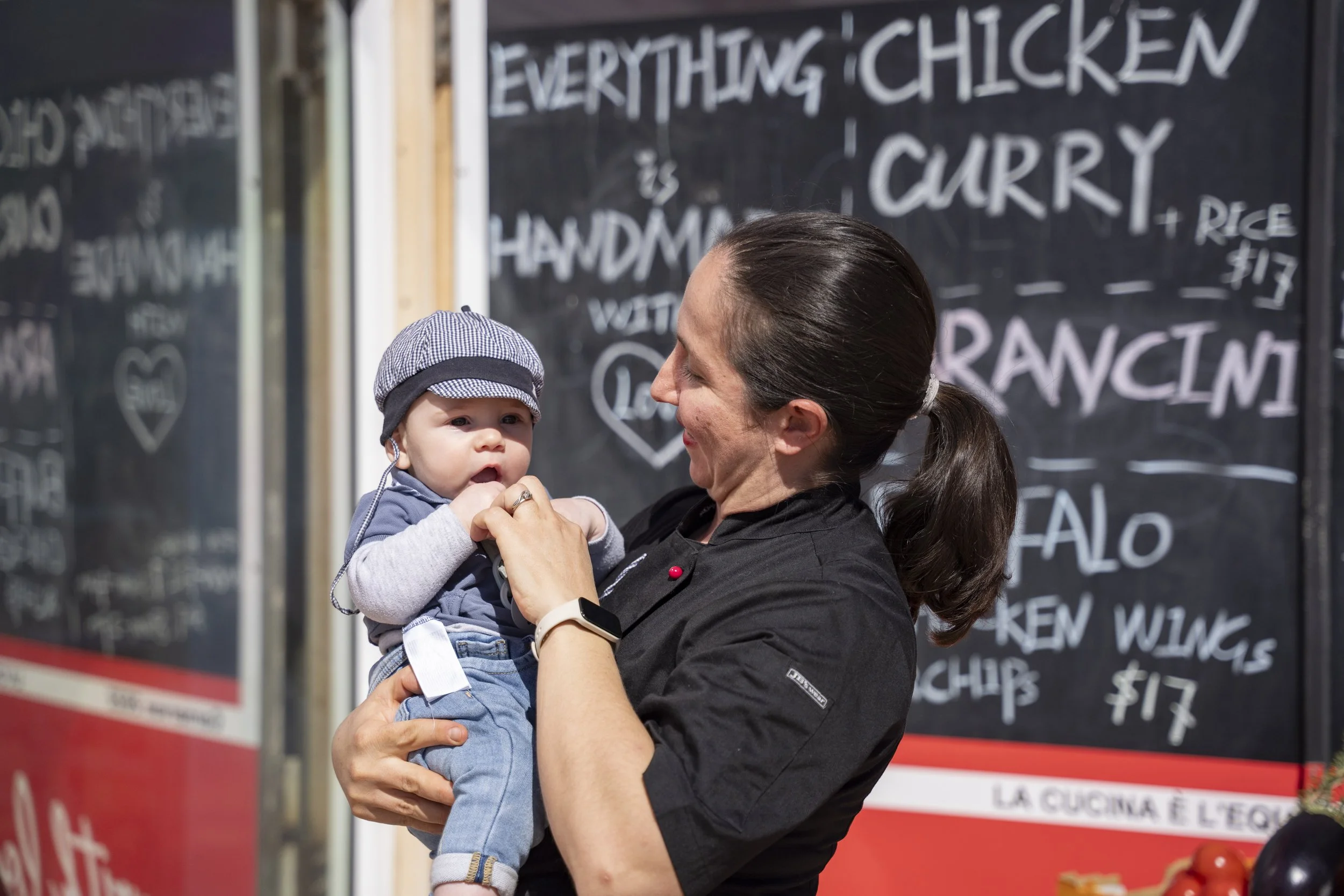 A woman in a black shirt holding a baby in front of a restaurant menu board.
