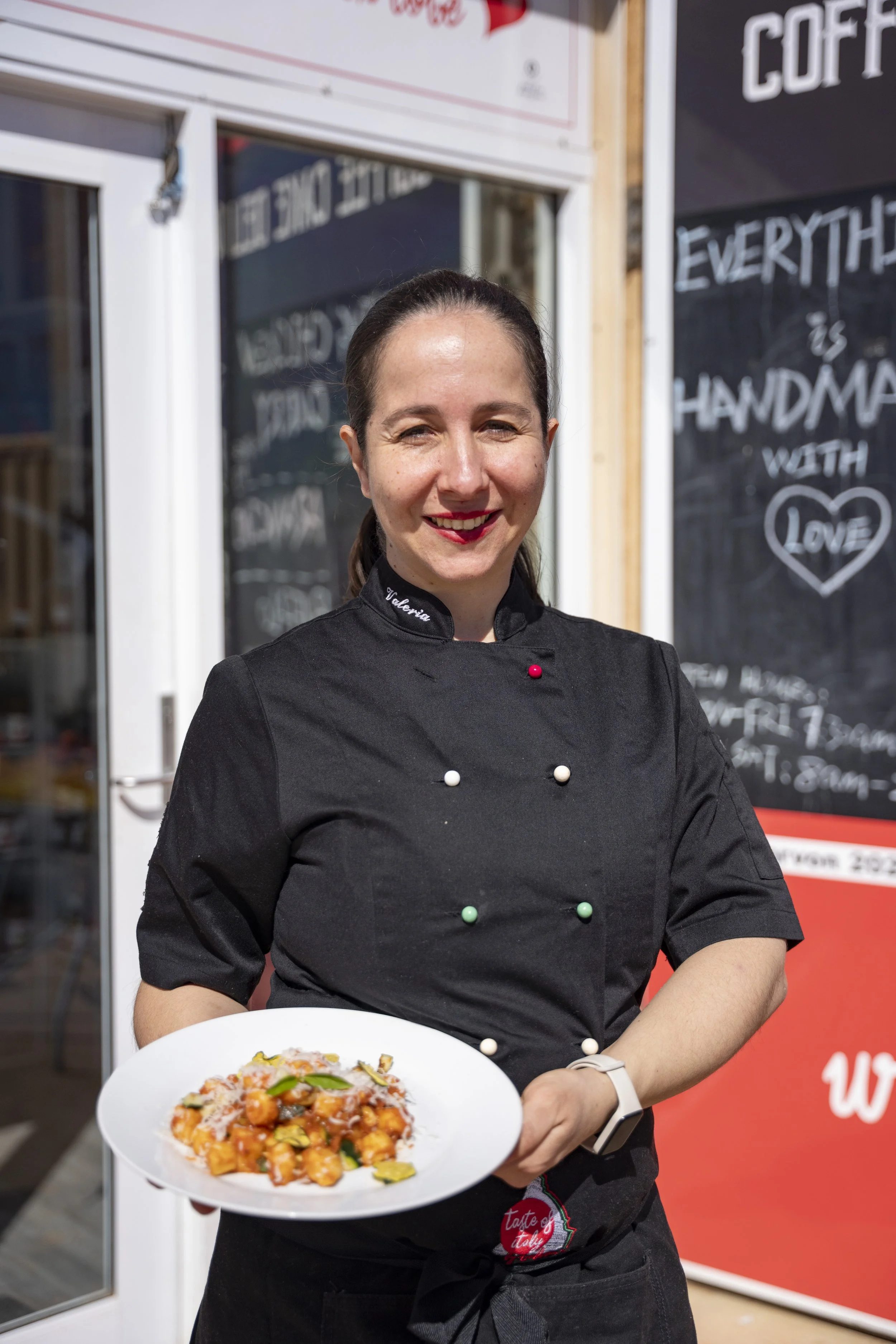 A woman in a black chef's coat holding a white plate with a colorful pasta dish outside a restaurant, smiling at the camera.