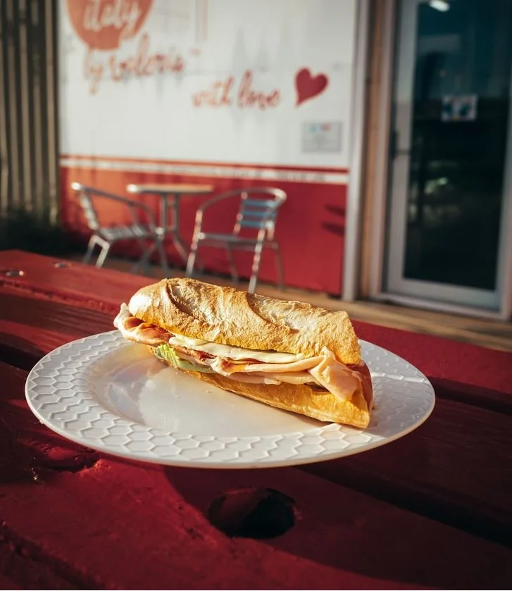 A ham and turkey sandwich on a white textured plate outside on a wooden table. In the background, there's a red and white building with two metal outdoor chairs.