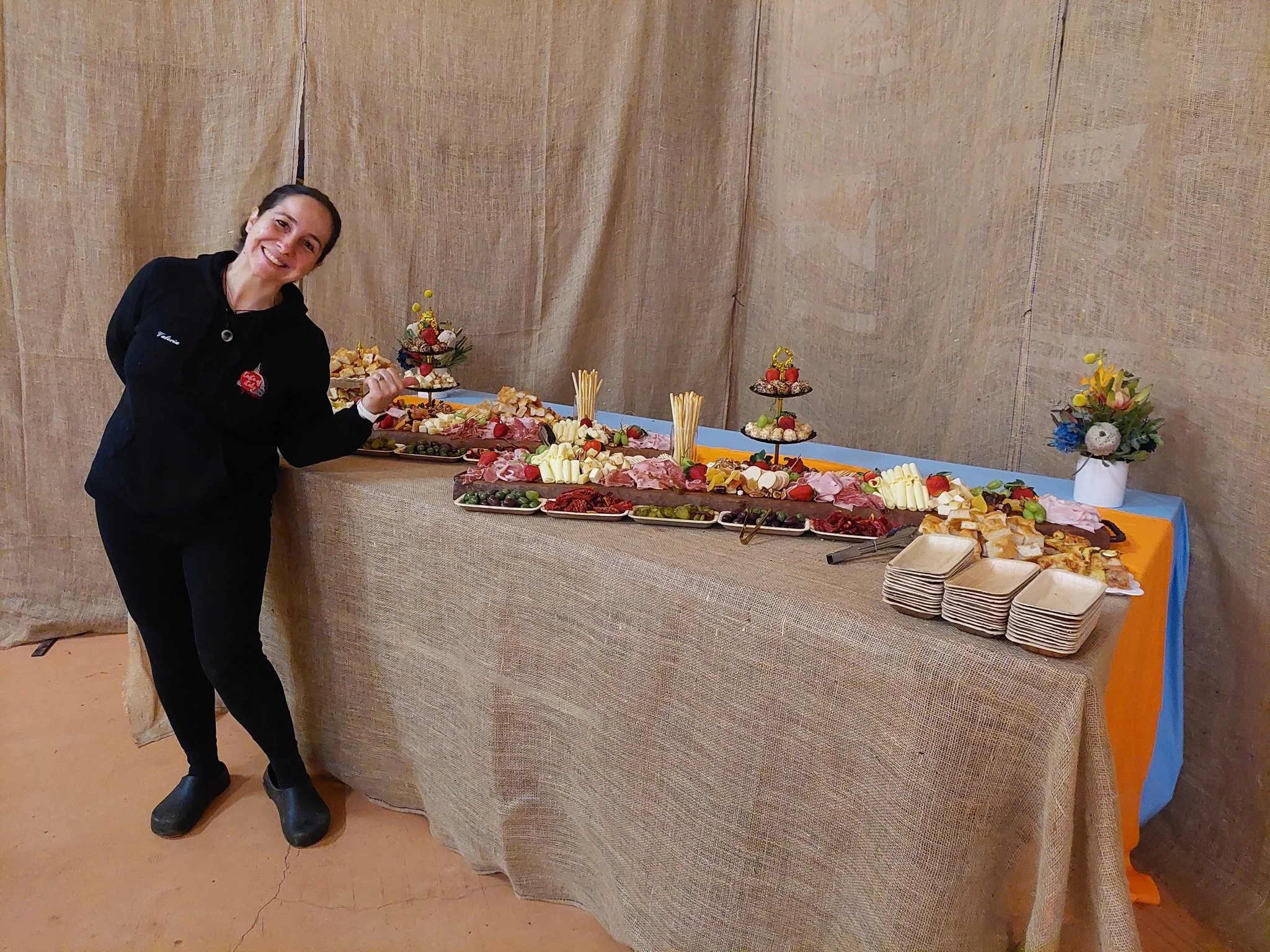 Smiling woman in black standing next to a long table of assorted cheeses, meats, fruits, and snacks, with decorative flower arrangements in the background.