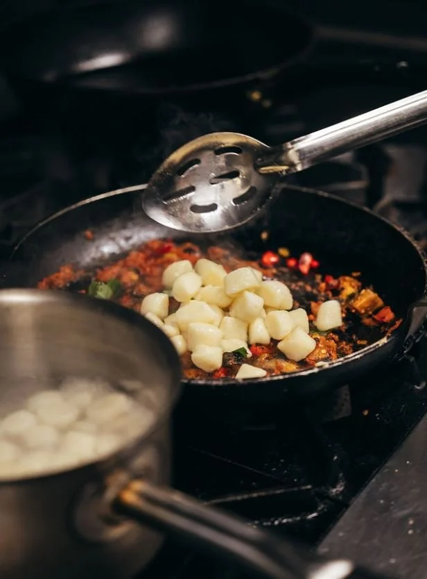 A frying pan on a stove with garlic and chili peppers cooking, and a slotted spoon resting on the pan. A pot of water is nearby.