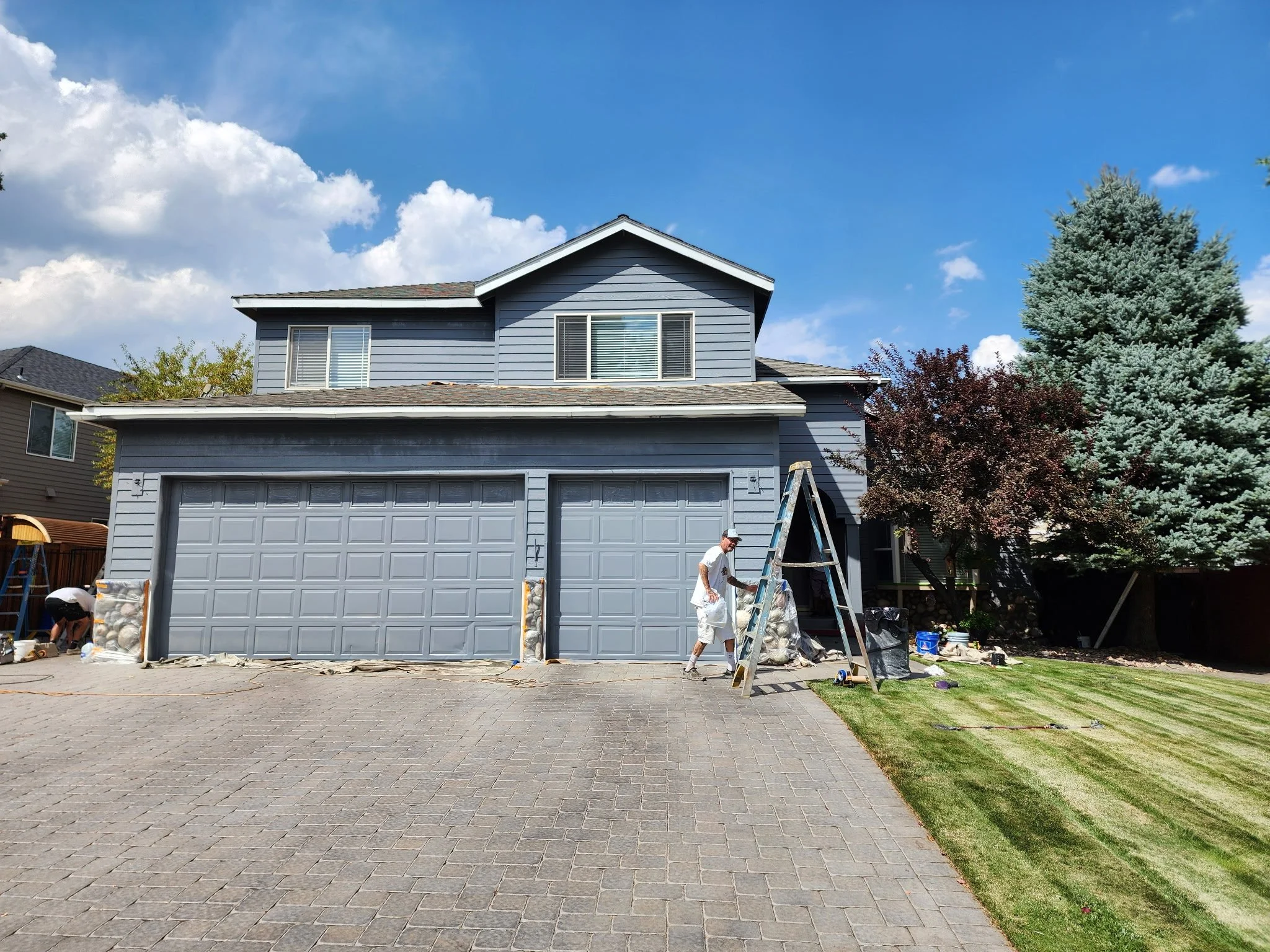 A two-story house with gray siding and a closed garage door is surrounded by a paved driveway and a front yard with some grass and plants. There are two workers in white shirts working on the house, one with a ladder. The house has two windows on the